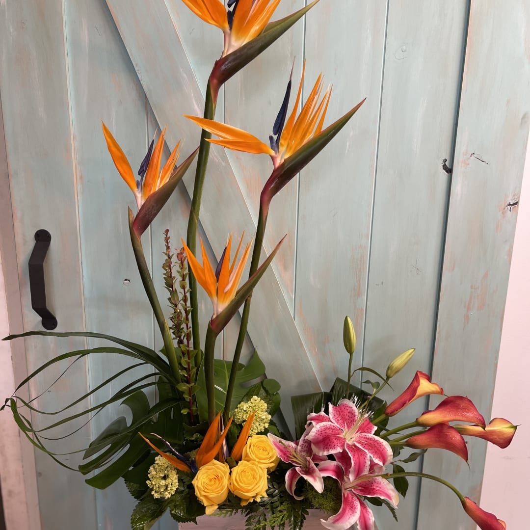 Tall tropical arrangement with orange bird-of-paradise, pink lilies, and yellow roses in a low container.