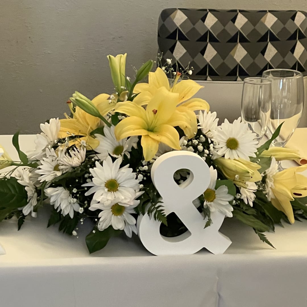Yellow lilies and white daisies arranged on a table with a white ampersand sign
