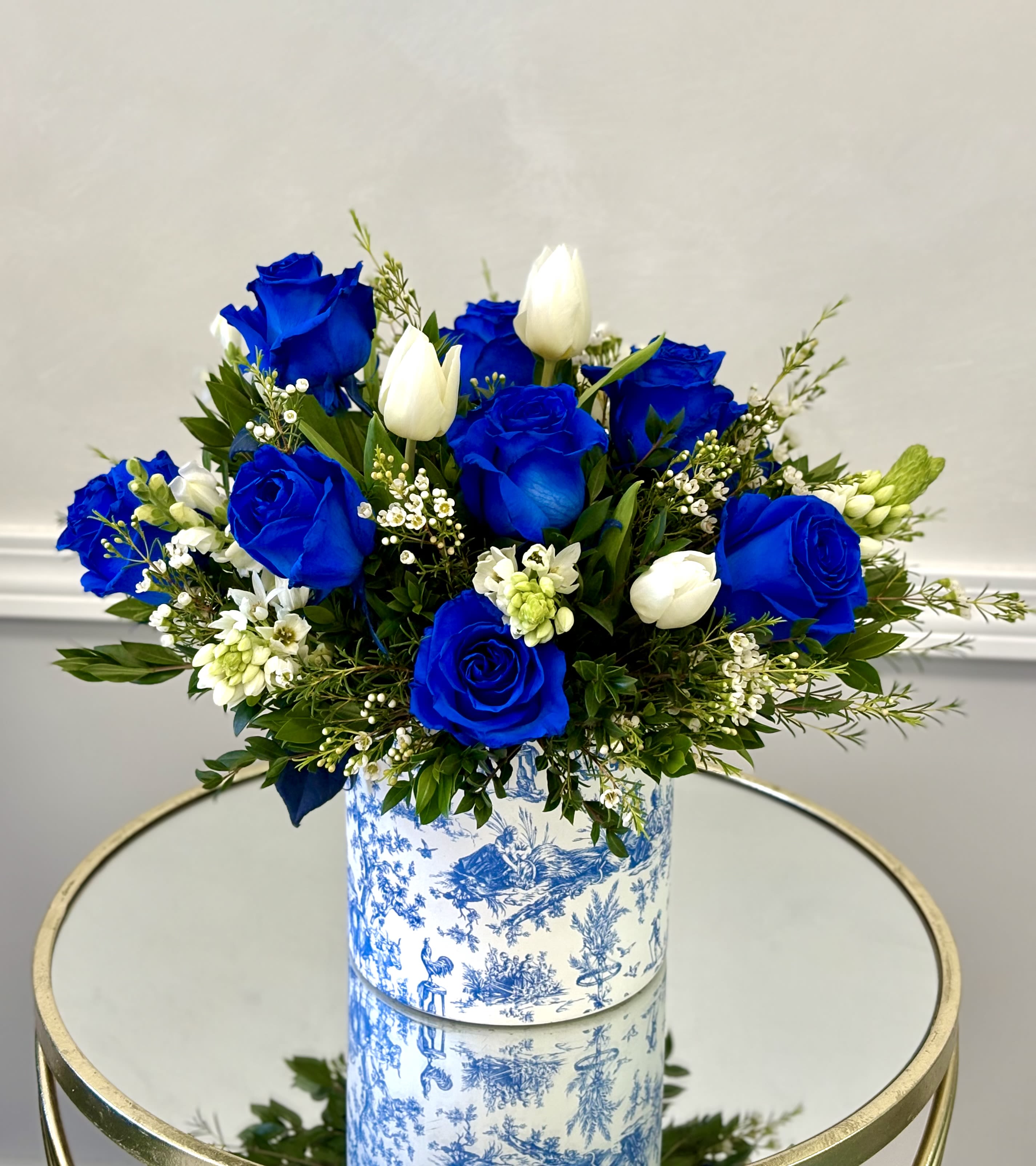 Royal Blue By VLM Flowers - Vivid royal-blue roses take center stage in this blue-and-white ceramic vase, creating a bold birthday bouquet or anniversary arrangement that feels both celebratory and refined. Paired with crisp white tulips and delicate white filler blooms, the contrast of blue, white, and fresh greenery gives the recipient an immediate sense of elegance - the kind of gift that makes someone feel truly honored and remembered.  The arrangement is gathered in a rounded, full design that sits in a patterned blue-and-white ceramic vase, offering a polished display straight from delivery to tabletop. Textures range from the velvety rose petals to the smooth tulip stems and airy filler blossoms, while the greenery frames each bloom for a balanced, long-lasting look. This is an excellent choice for birthdays, anniversaries, or a thoughtful thank-you that needs to read as distinctive and memorable.  Designed to arrive ready-to-enjoy, the ceramic vase adds instant presence and makes the bouquet easy to place in an office or home. The colorway and composition suit recipients who appreciate sophisticated, cool-toned arrangements - someone who will feel seen and celebrated by this striking, elegant display.