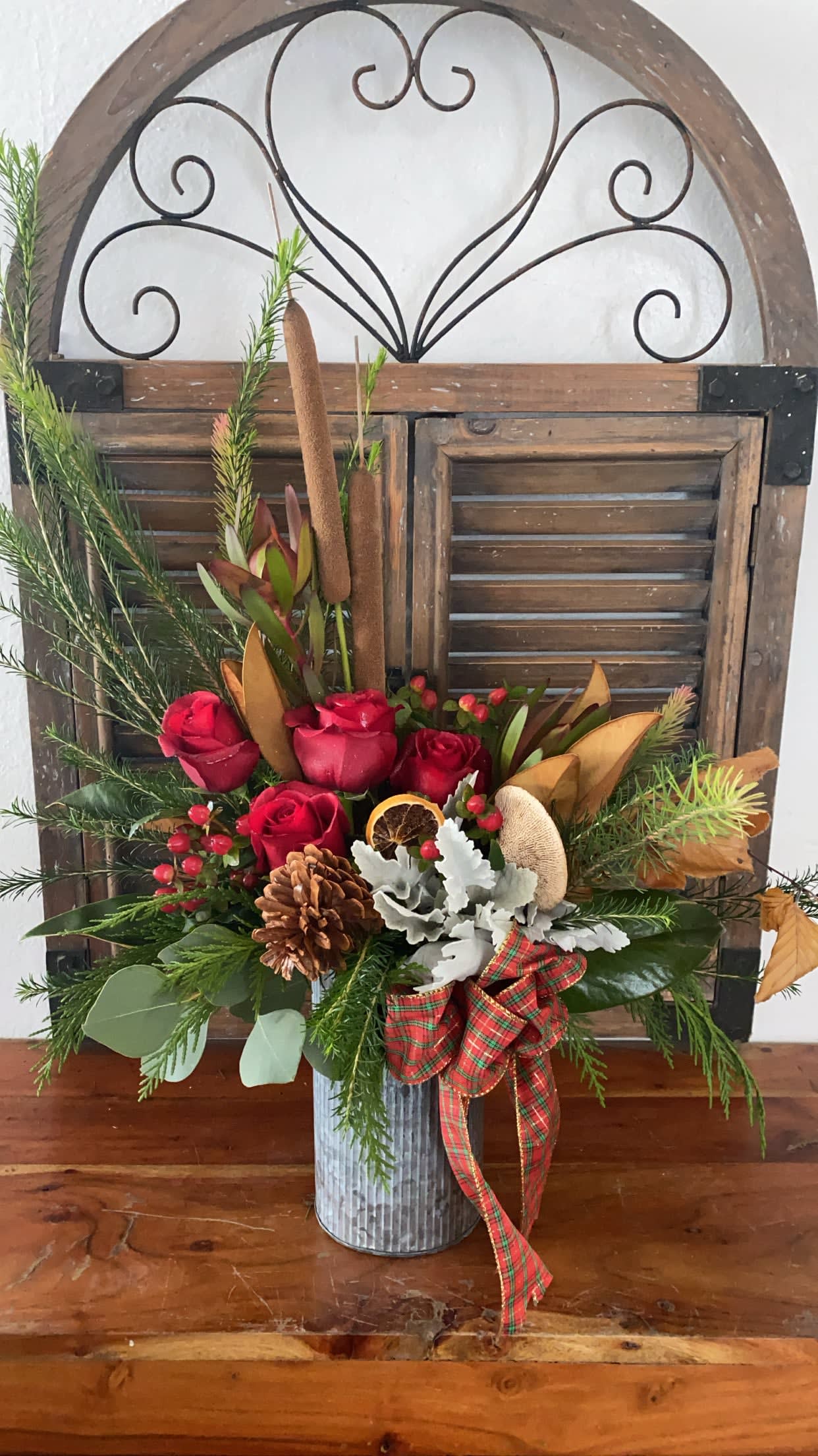 Red roses in a metal vase with pine, pinecones, and a plaid ribbon