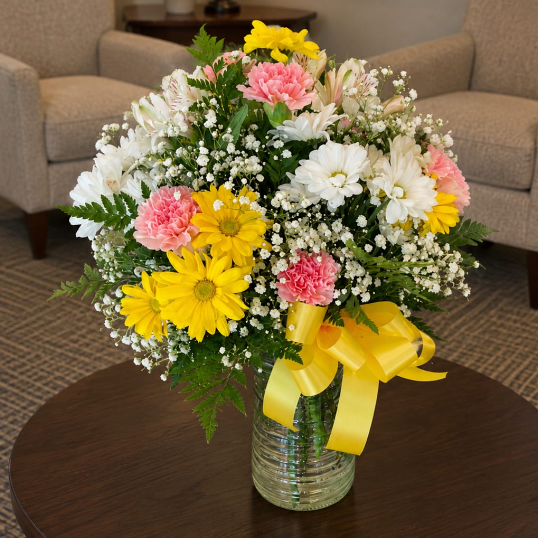 Mixed bouquet of daisies, carnations, and baby's breath in a glass vase with a yellow ribbon