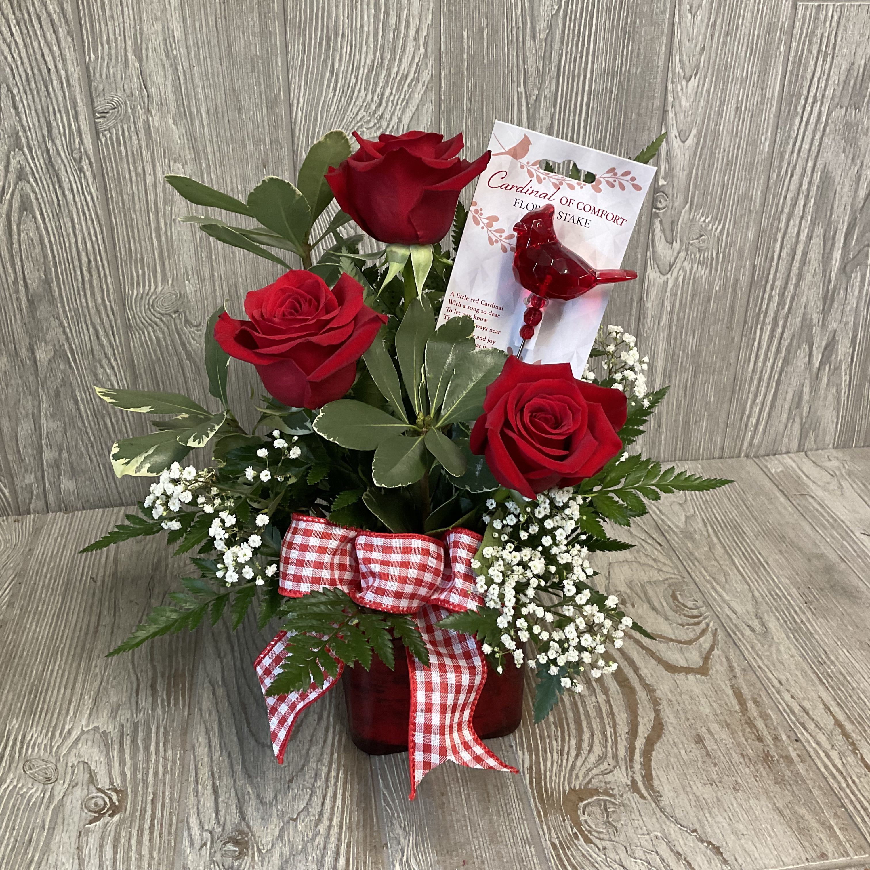 Cardinal of Comfort  - Three deep red roses rise from a compact cluster of delicate white filler blooms, creating a warm, heartfelt arrangement perfect for sympathy, a funeral, or a gentle "thinking of you" message. The upright composition and the saturated red glass vase give the bouquet a comforting presence-the kind of arrangement that reads as both reverent and tender when you want to offer support or remembrance.  The three roses are accented by soft white sprigs that add lightness and texture, tied together with a red-and-white gingham ribbon bow that introduces a cozy, homespun note. A small red cardinal figurine on a floral stake, complete with a printed card, nestles among the flowers as a sentimental touch suggesting hope and enduring memory. The design is compact and upright, making it suitable for placement on a mantel, bedside table, or small memorial display.  Thoughtful for a family member, friend, or neighbor who needs comfort, this arrangement sends a clear message of care and remembrance without overwhelming the space. Vase and accents are included; colors and natural flowers may vary slightly with season and availability, but the cardinal pick and gingham bow remain as the arrangement's signature elements.