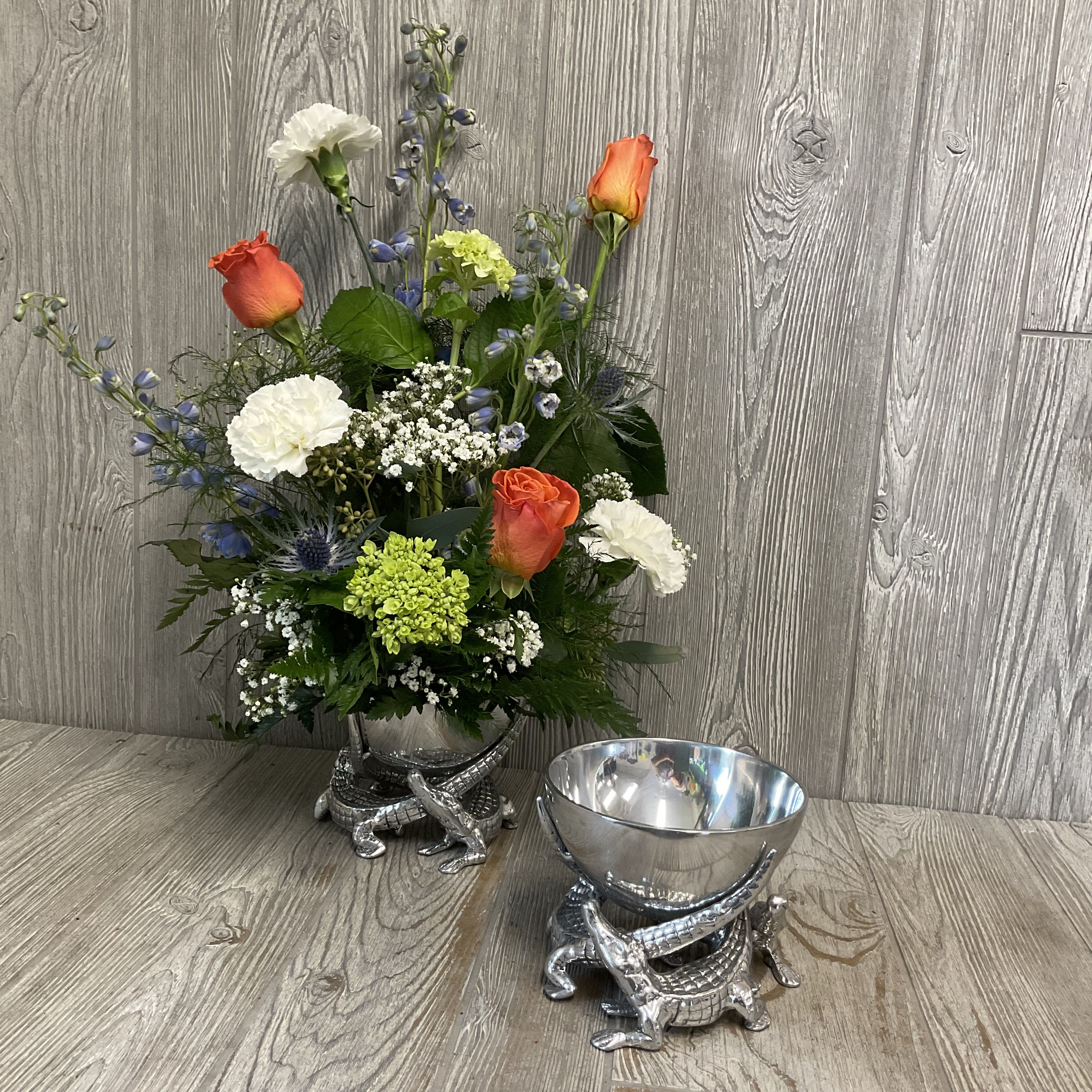 Gator Pedestal Arrangement by Arthur Court - Bright orange roses set against soft blue delphinium, crisp white carnations, and a vivid green hydrangea cluster create a cheerful, sculptural centerpiece-perfect as a birthday bouquet, congratulations arrangement, or a playful decorative accent for the home. The rounded, medium-height floral shape pops from a shiny pewter metal bowl that's cradled by 3 detailed alligator figures, giving the arrangement a whimsical, modern character that will make the recipient feel truly celebrated.  Flowers are arranged with small filler blooms for texture and balance, mixing warm orange, cool blue and white tones for lively contrast. The offering includes the distinctive silver metal bowl, by Arthur Court, on a sculpted alligator base. A piece that works as striking serveware or display objects. Each set arrives in a light blue Arthur Court gift box, ready for gifting-an ideal choice when you want to send something stylish, unexpected, and memorable.