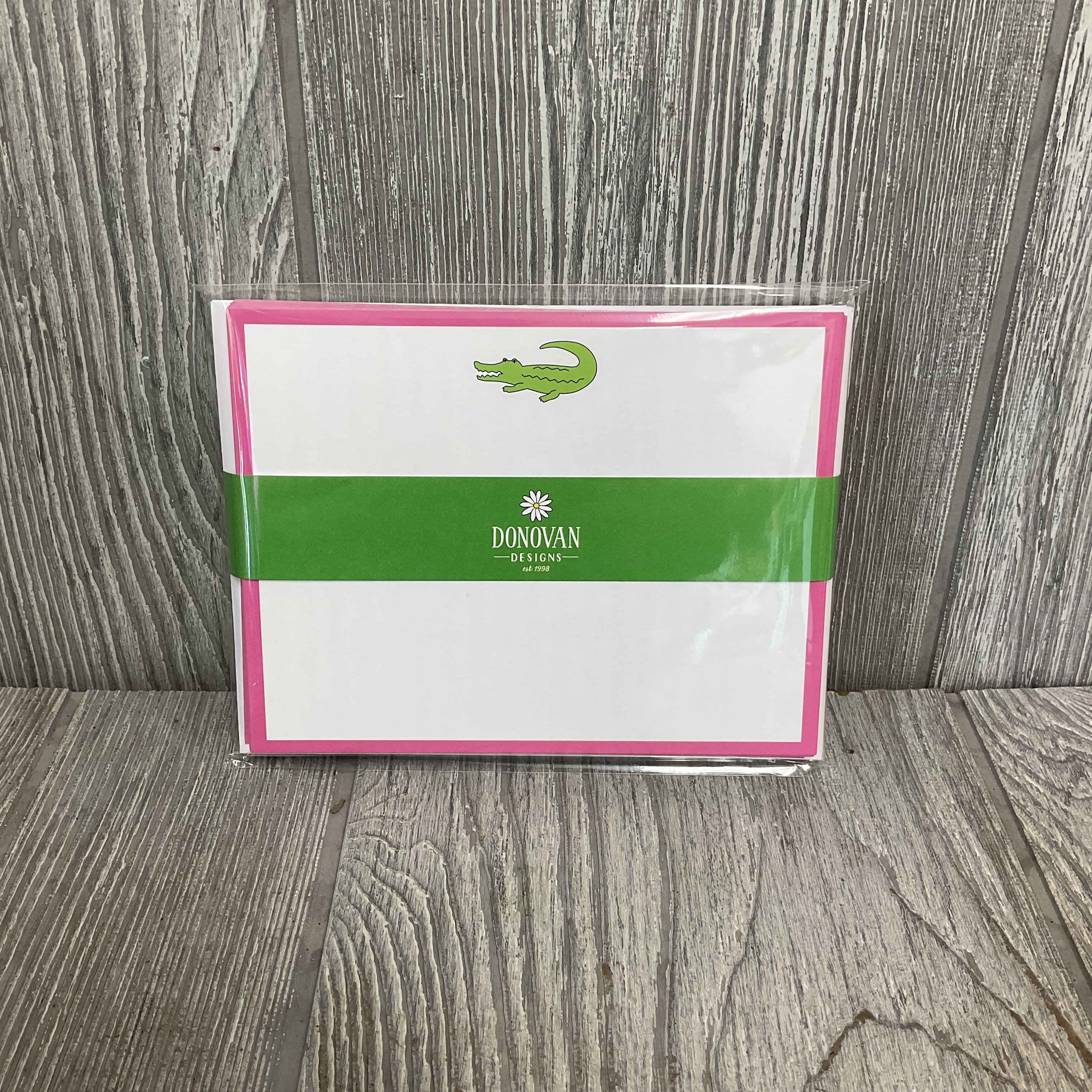 Gator Flat Notecards with Envelopes by Donovan Designs - Bright, playful notecards perfect for everyday messages and casual gifting - these flat stationery cards pair a bold pink border with a cheerful green alligator motif for a fresh, upbeat look. Use them as birthday notes, party thank-yous, or simple just-because messages when you want the recipient to smile; the design reads youthful and friendly rather than formal, so they're ideal for kids, friends, or anyone who appreciates a whimsical touch.  Each set includes 10 flat greeting cards with coordinating white envelopes, packaged in clear plastic for a retail-ready presentation. The card front shows a clean white face framed in bright pink with a small green alligator near the top; the back features pink-and-white vertical stripes with repeated alligator prints. Lightweight and easy to mail or tuck into a bouquet, these notecards add a playful accent to gifts and arrangements.
