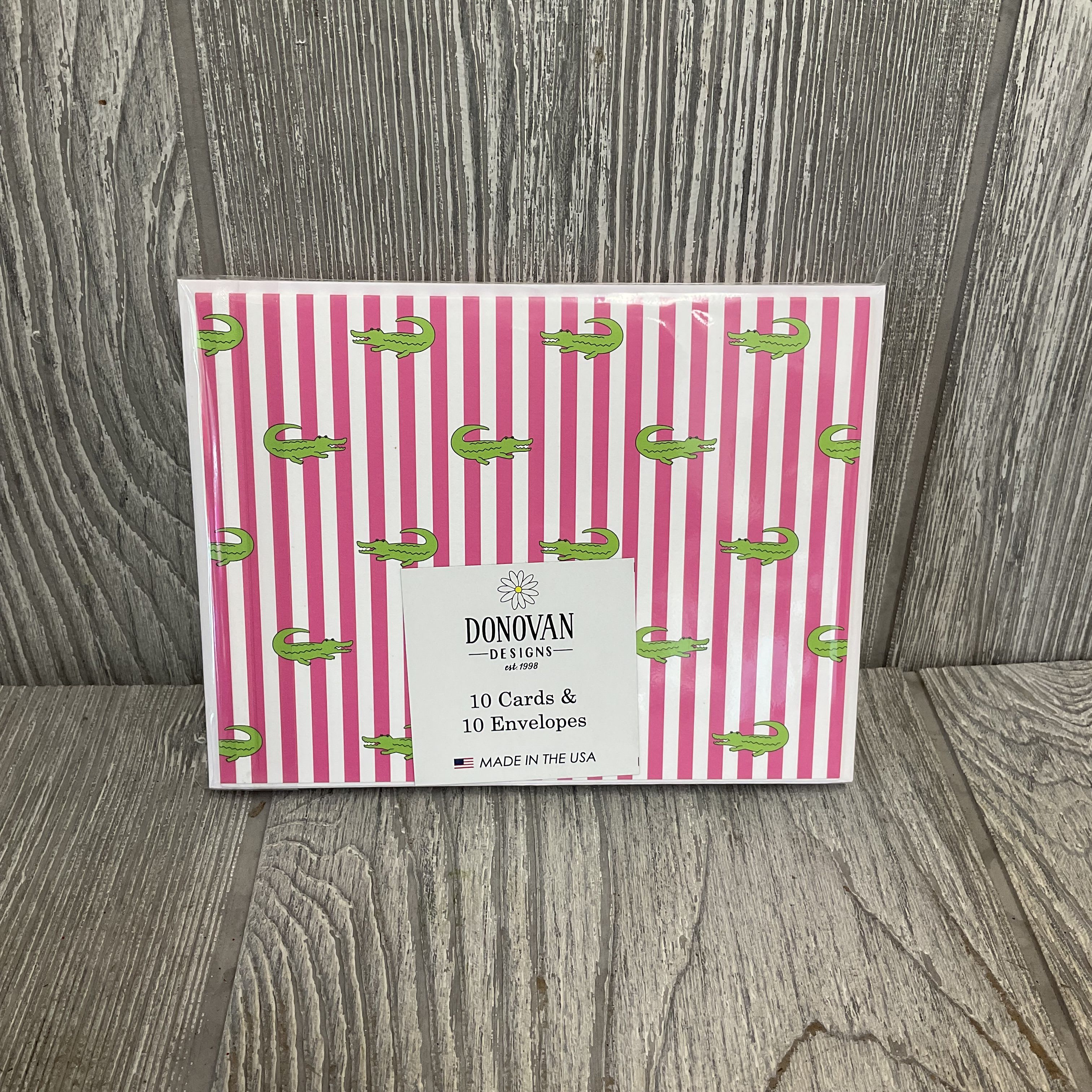 Gator themed Folding Cards by Donovan Designs - Bright pink-and-white vertical stripes meet cute green alligators in a boxed set of Gator-themed folded greeting cards-perfect for birthday notes, new-baby congratulations, or just-because surprises. These playful greeting cards and envelopes read as cheerful and casual, the kind of stationery that makes the person you're thinking of smile before they even open it. The printed cards' whimsical illustrations and friendly color palette make them a great match for lighthearted celebrations and gift accompaniments.  Each set includes 10 printed folded greeting cards, matching envelopes, and retail-ready packaging with a simple white label; each card has a plain white inside and is printed with an outside of pink and white stripes and cute gator print. These cards pair especially well with baby gifts, cheerful bouquets, or party favors.