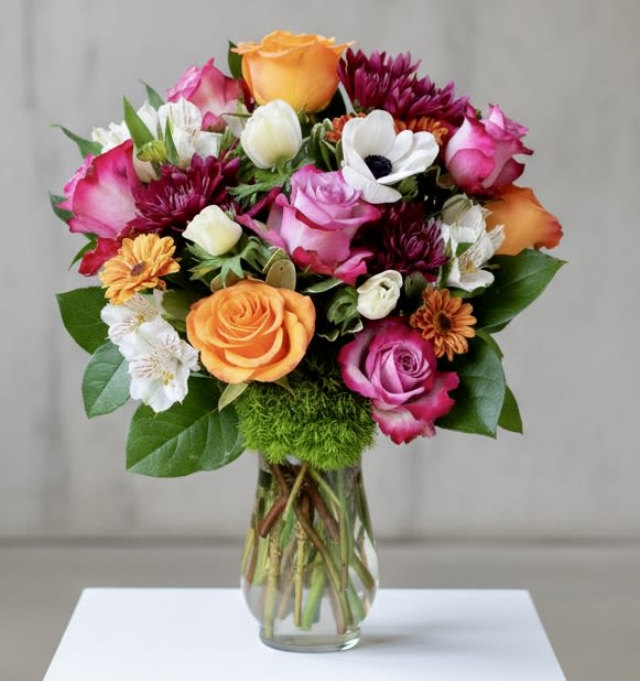 Birthday Blossoms  - A cheerful, color-rich birthday bouquet that makes the person you're thinking of feel genuinely celebrated: dense clusters of orange and hot-pink-edged roses mingle with striking white anemones (dark centers) to create an immediate focal pop. This flower arrangement for birthdays also works beautifully for get well wishes, graduations, thank-you gestures, or just-because moments-anytime you want to lift someone's spirits with bold, joyful color.  Featuring: Ecuadorian Roses, French Dianthus, White Himalaya Alstroemeria, Colorful Spray Mums, Panda Anemones, Israeli Ruscus and Lemon Leaf.  Presentation Options: Comes in a Clear Glass Vase. Upgrade to a Gold Ceramic Vase for $22 OR at Checkout, under "Instructions" request a Hand Tie Bouquet (delicately wrapped in paper with a bow) or Low Centerpiece at no extra cost.      Care tip: refresh the water and trim stems every few days and remove those blooms that expire first to keep the mixed blooms looking their best.  