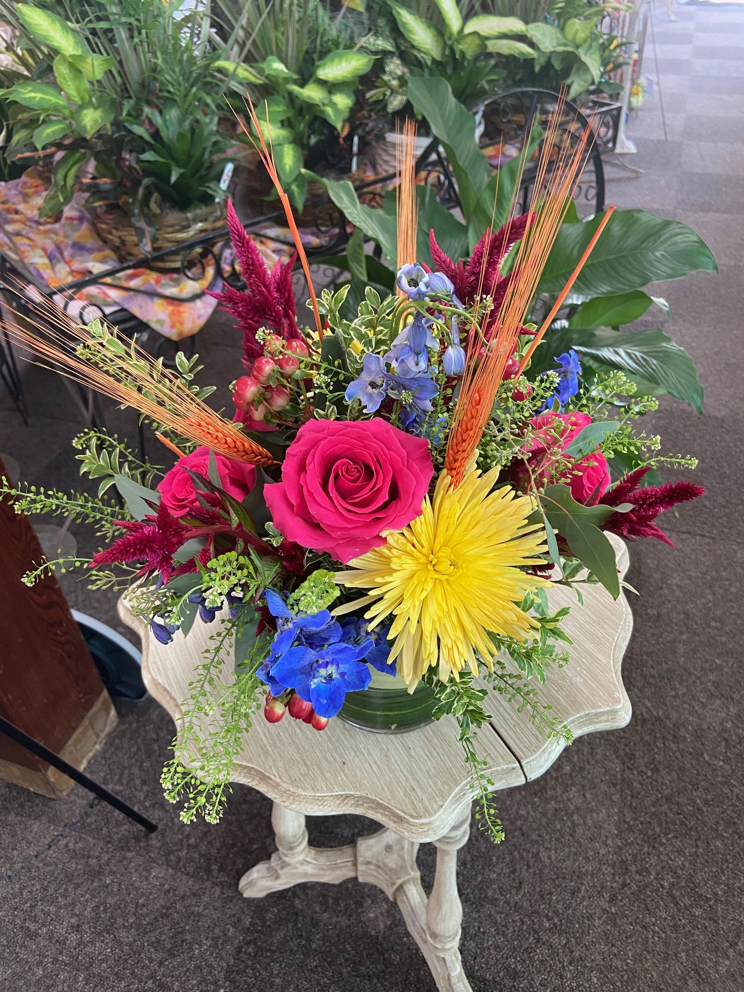 Bright mixed arrangement with hot pink rose, yellow mum, blue delphinium and wheat stems in a clear glass vase.