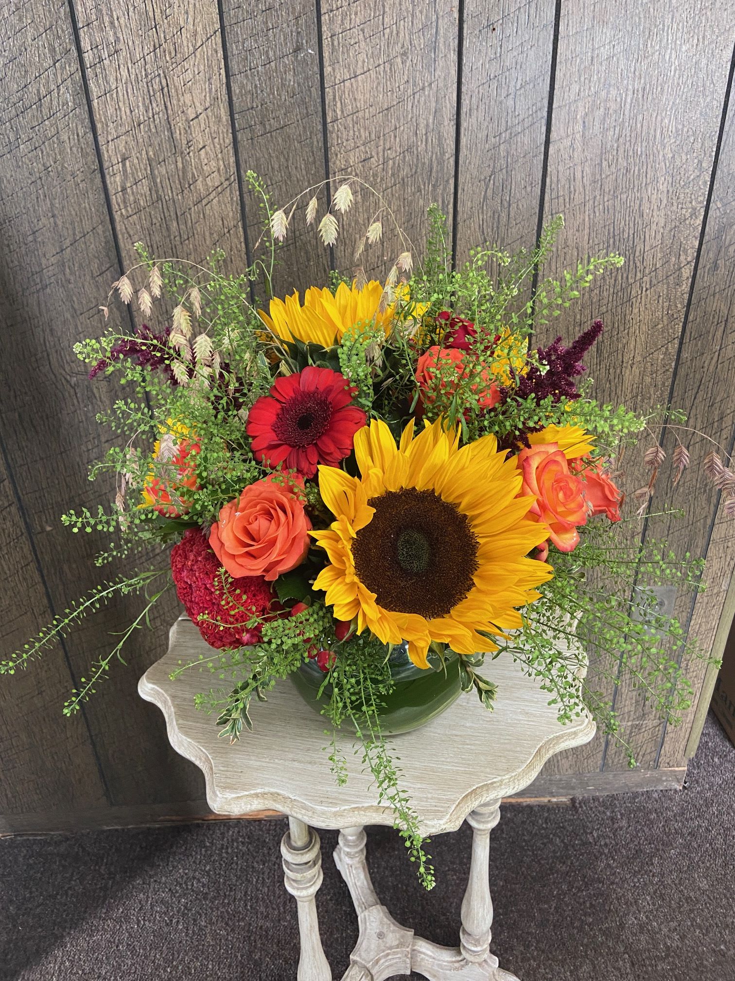 Low arrangement of sunflowers, orange roses, and red blooms in a green bowl on a small table