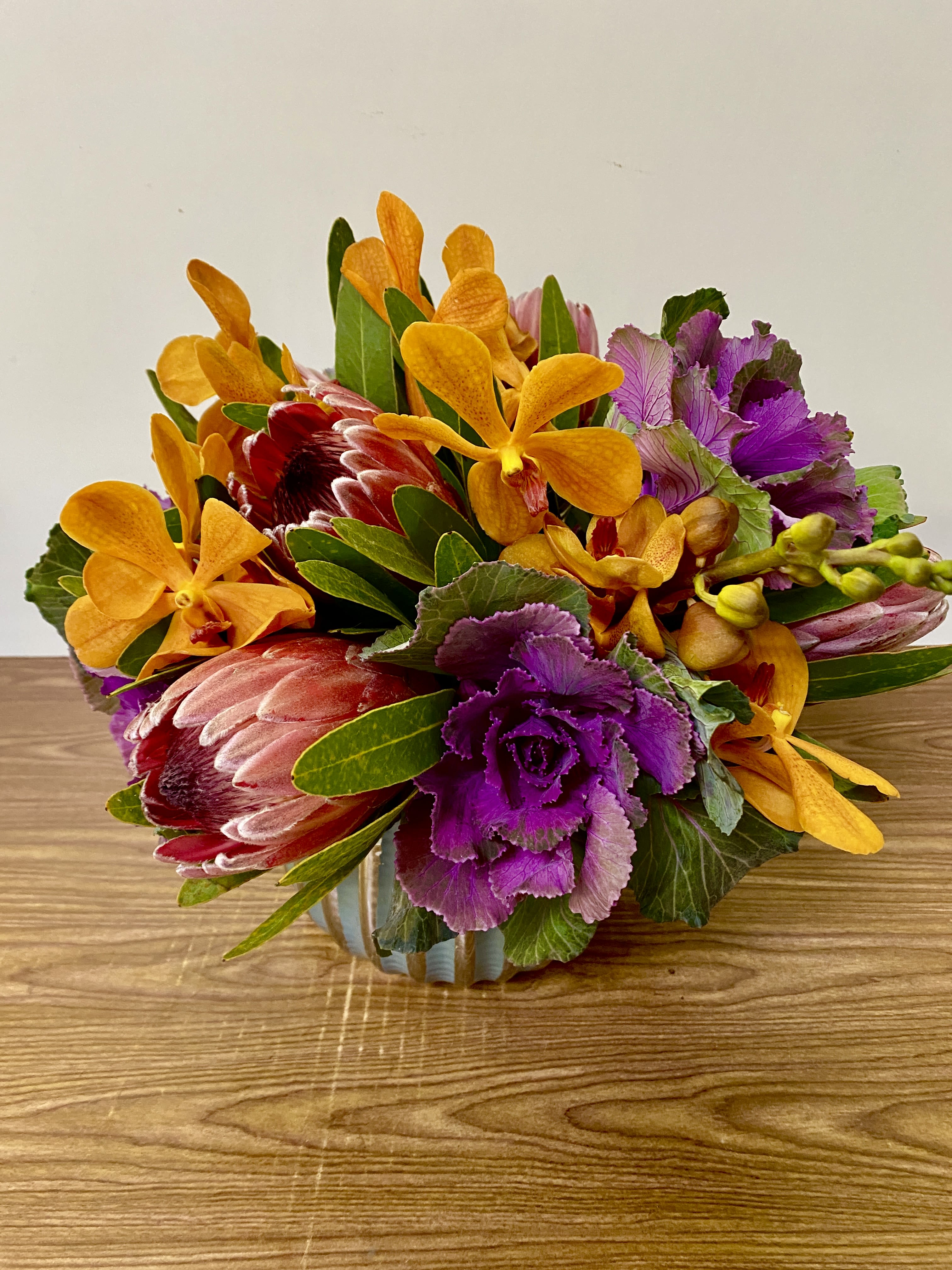 Mixed bouquet with orange orchids, purple kale, and red protea in a low container