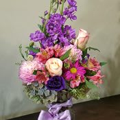 Tall mixed bouquet of purple and pink flowers with roses and daisies in a glass vase tied with a lavender bow