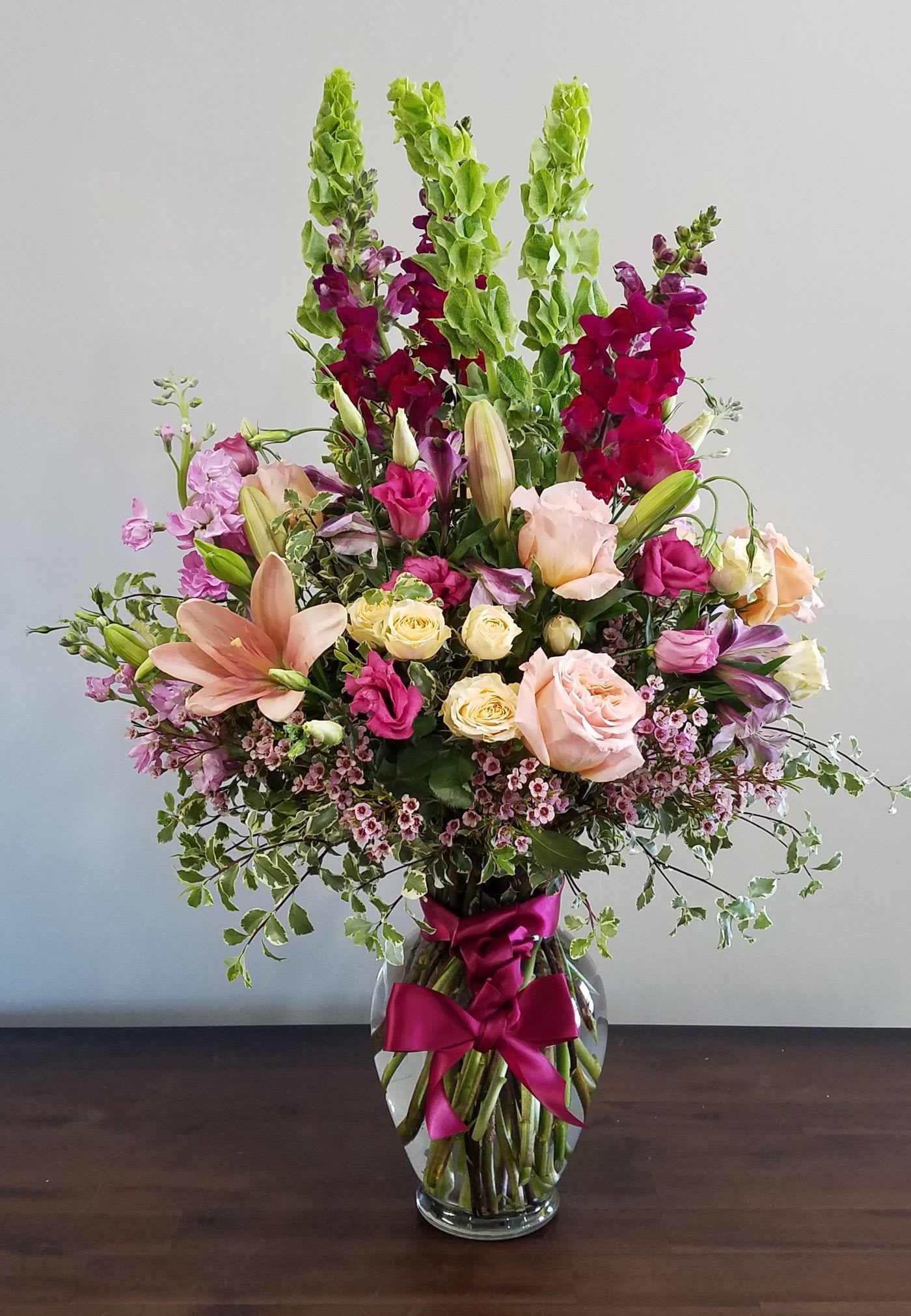 Tall mixed bouquet of pink, peach, and yellow flowers with green spikes in a clear vase with a magenta bow