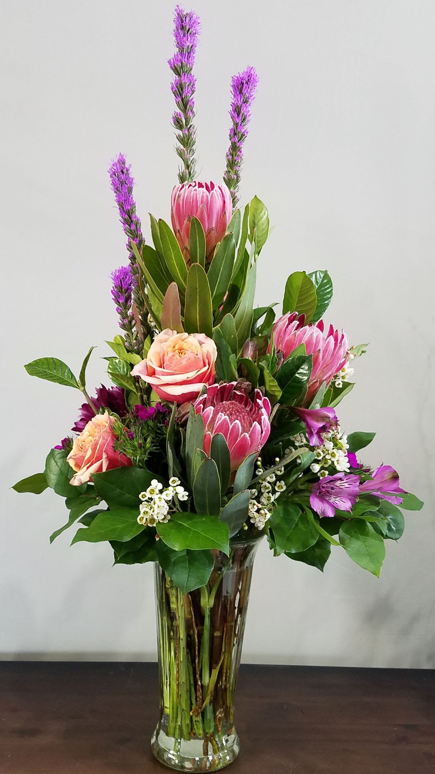 Tall arrangement of pink protea, roses, and purple flowers in a clear glass vase