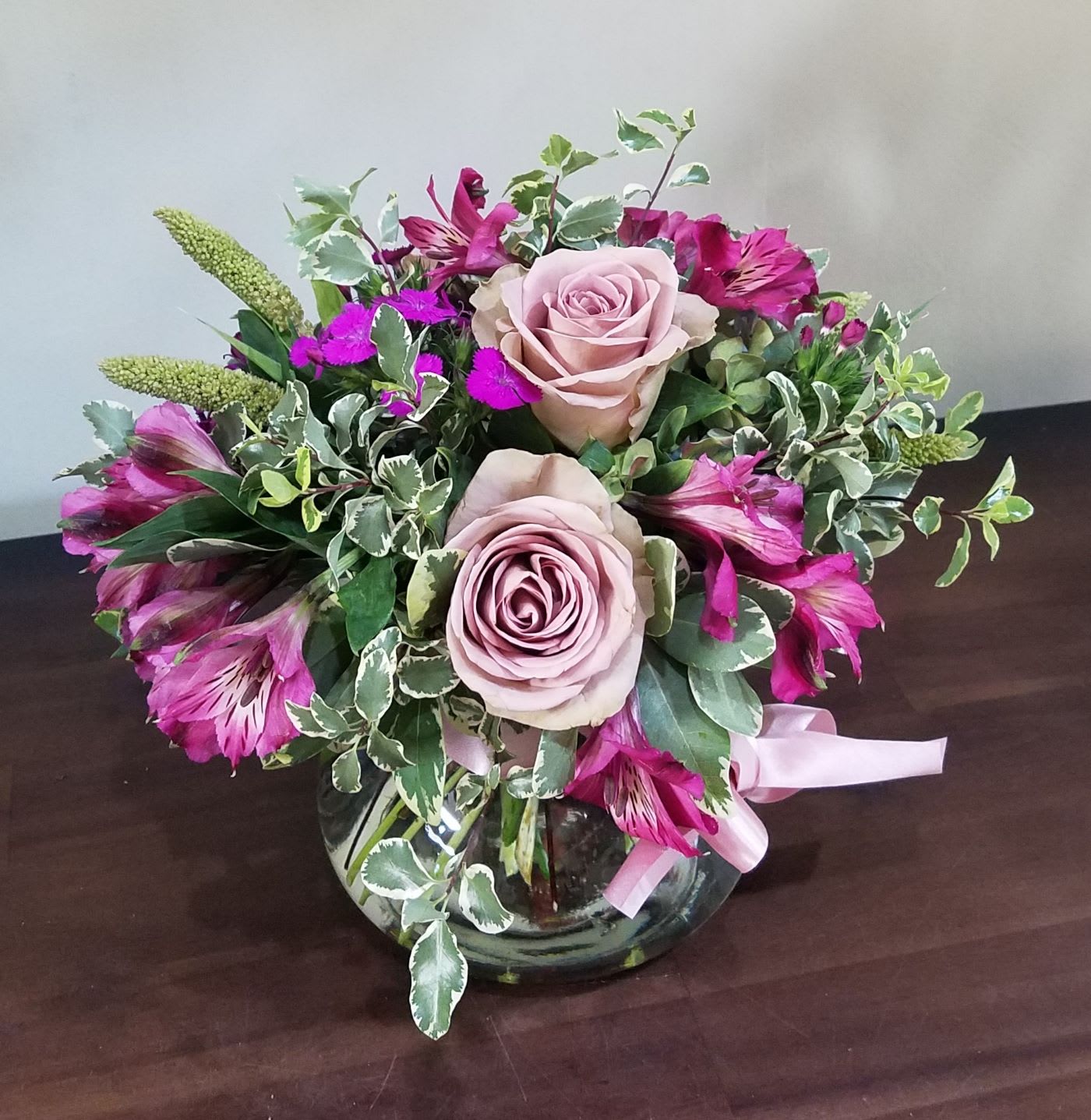Round bouquet of mauve roses and magenta alstroemeria in a clear glass bowl vase with pink ribbon