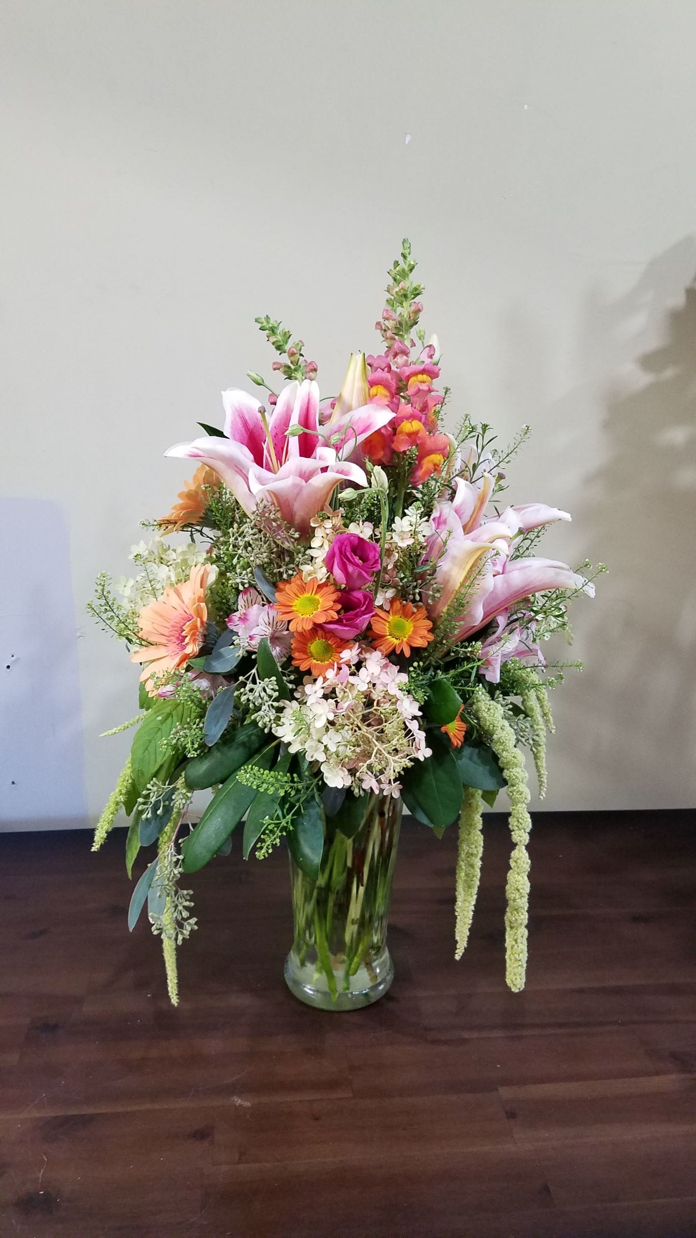 Tall mixed arrangement of pink lilies, orange daisies, and assorted flowers in a clear glass vase