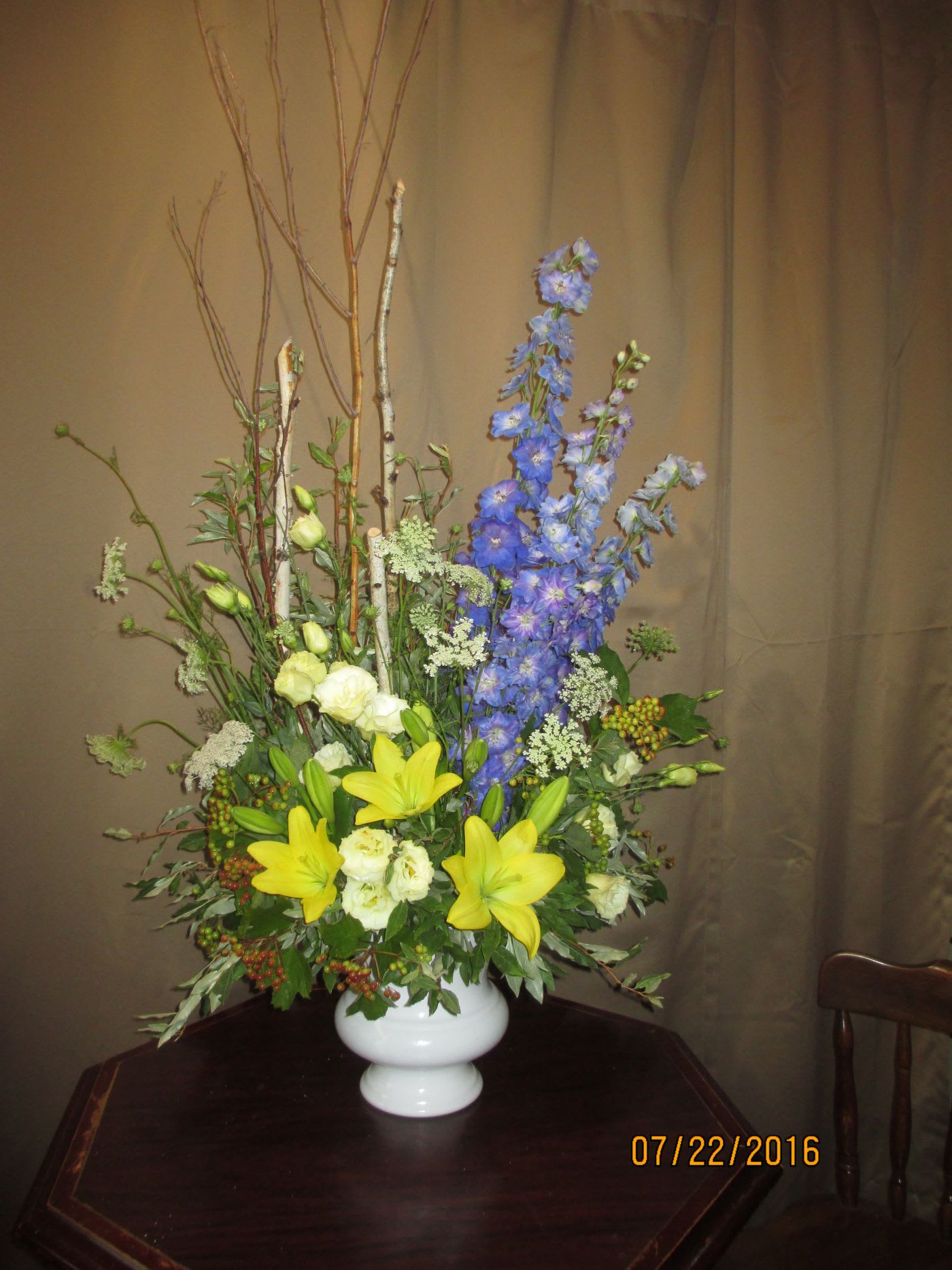 Tall arrangement of yellow lilies, white blooms, and blue delphinium in a white vase with branches