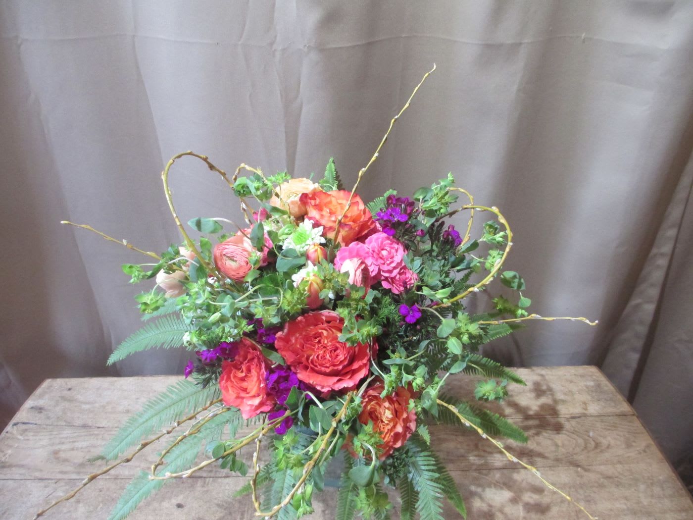 Colorful arrangement of orange and pink roses with purple flowers and curly branches in a low vase
