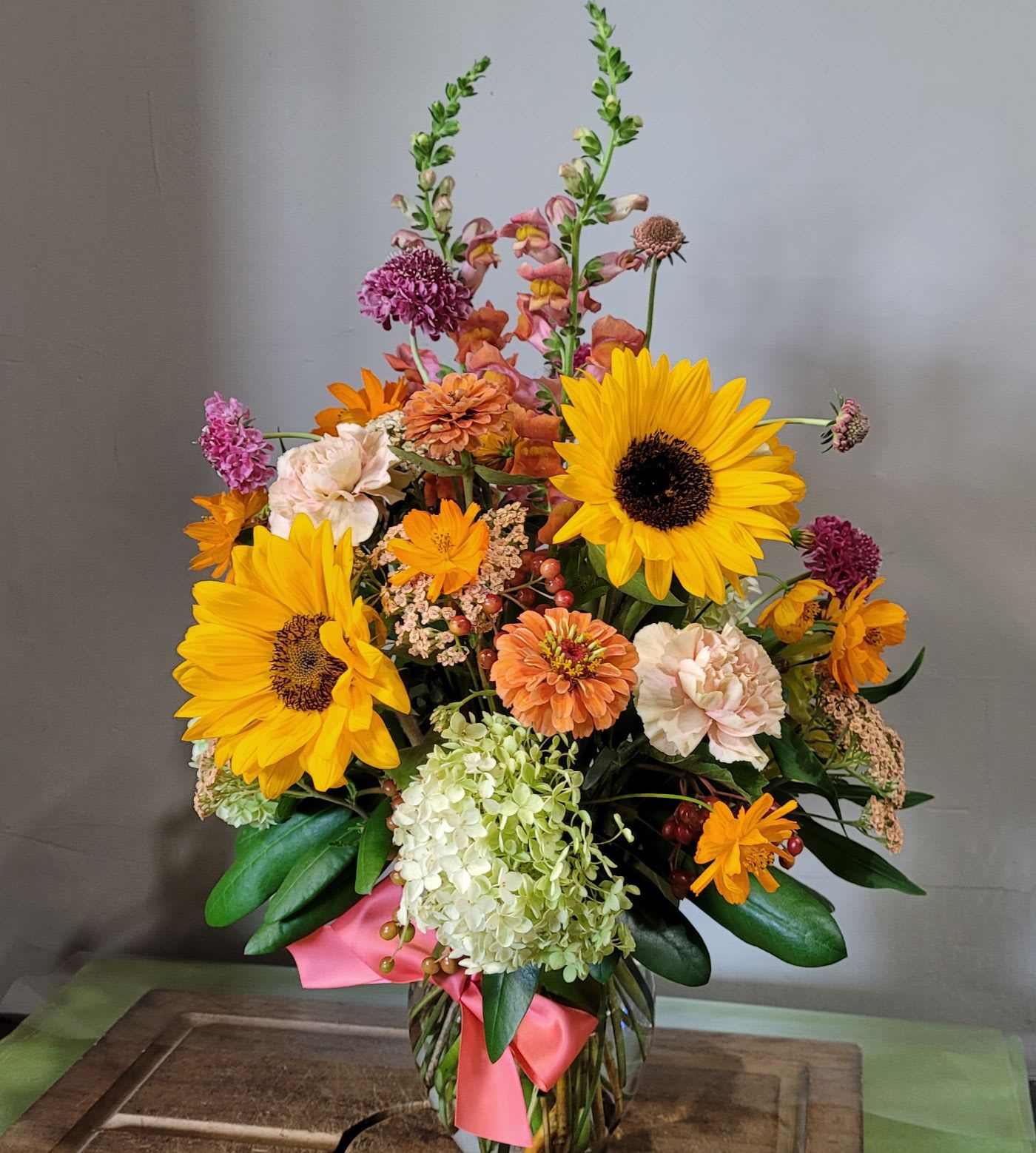 Tall mixed arrangement with sunflowers, hydrangea, and assorted orange and peach blooms in a glass vase.