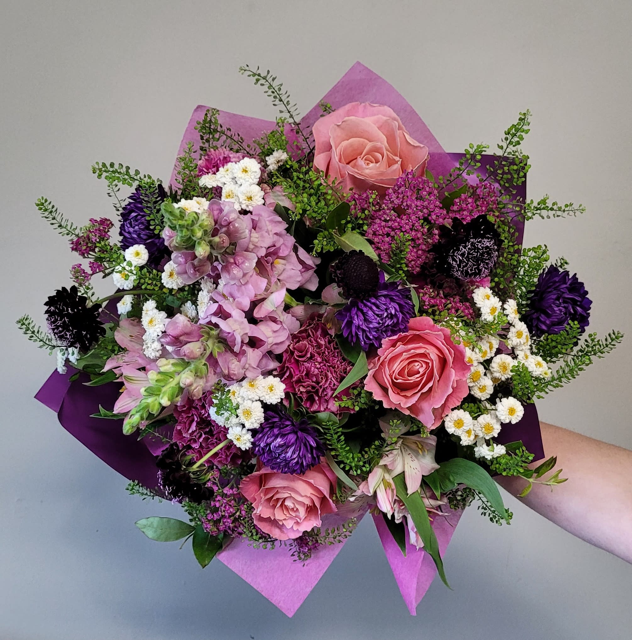 Hand-tied bouquet of pink roses, purple flowers, and white daisies wrapped in pink and purple paper