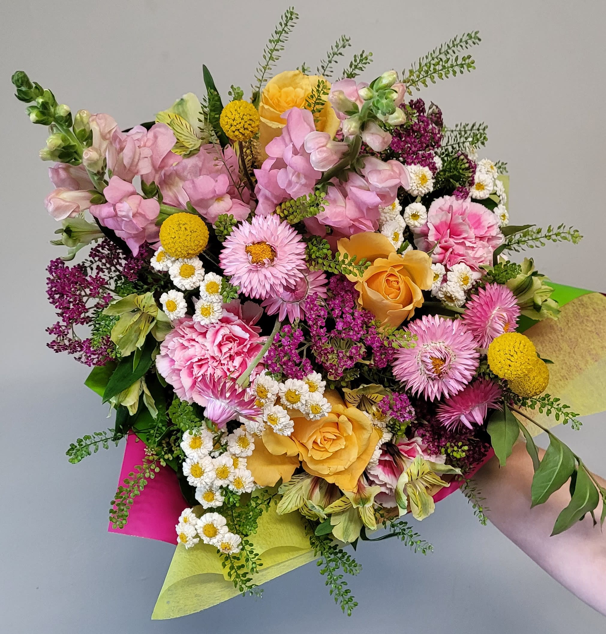 Hand-tied bouquet of pink and yellow flowers including roses, snapdragons, carnations, and daisies in bright wrap.