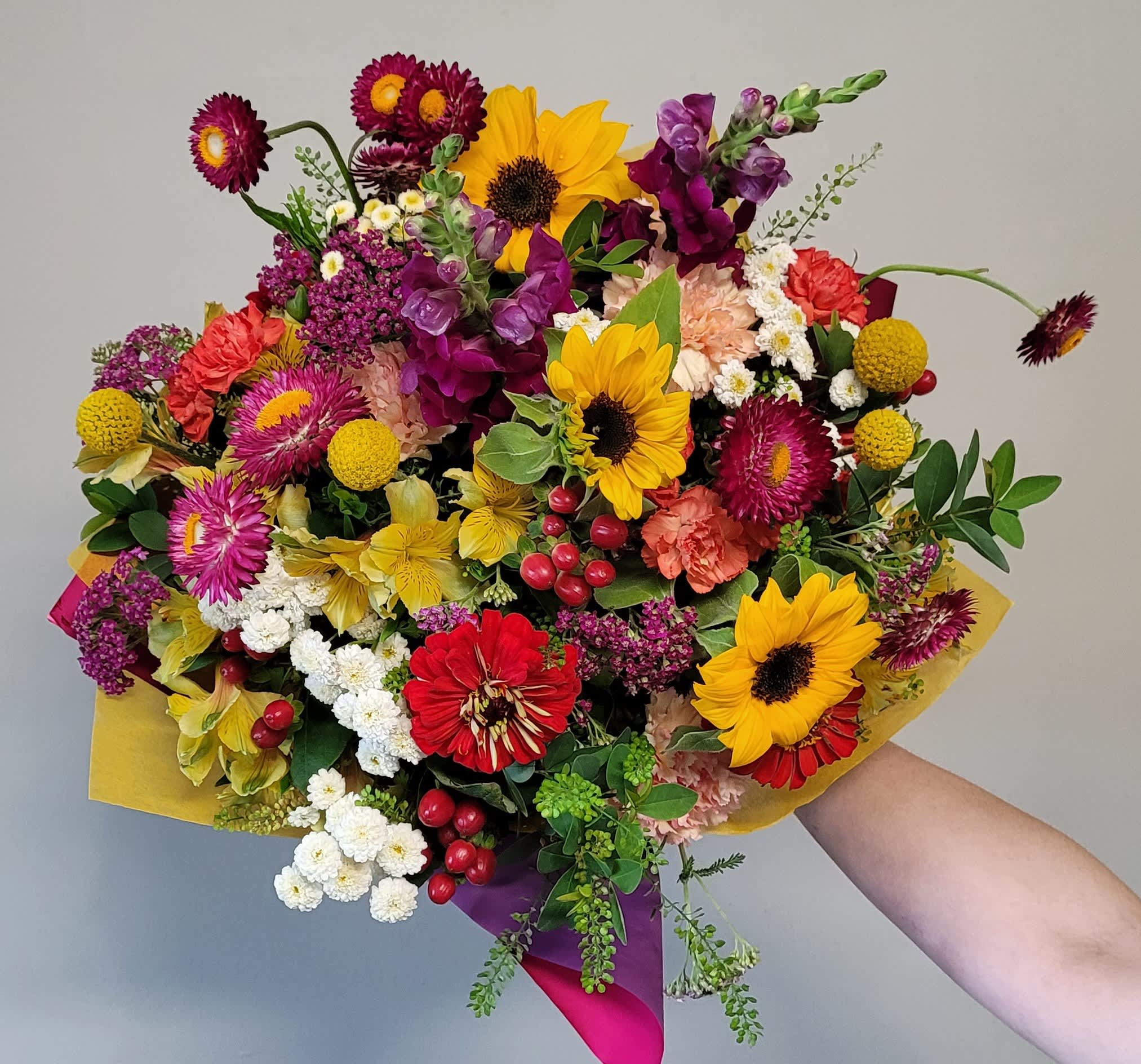 Hand-tied bouquet of bright sunflowers and mixed red and purple blooms wrapped in yellow and magenta paper