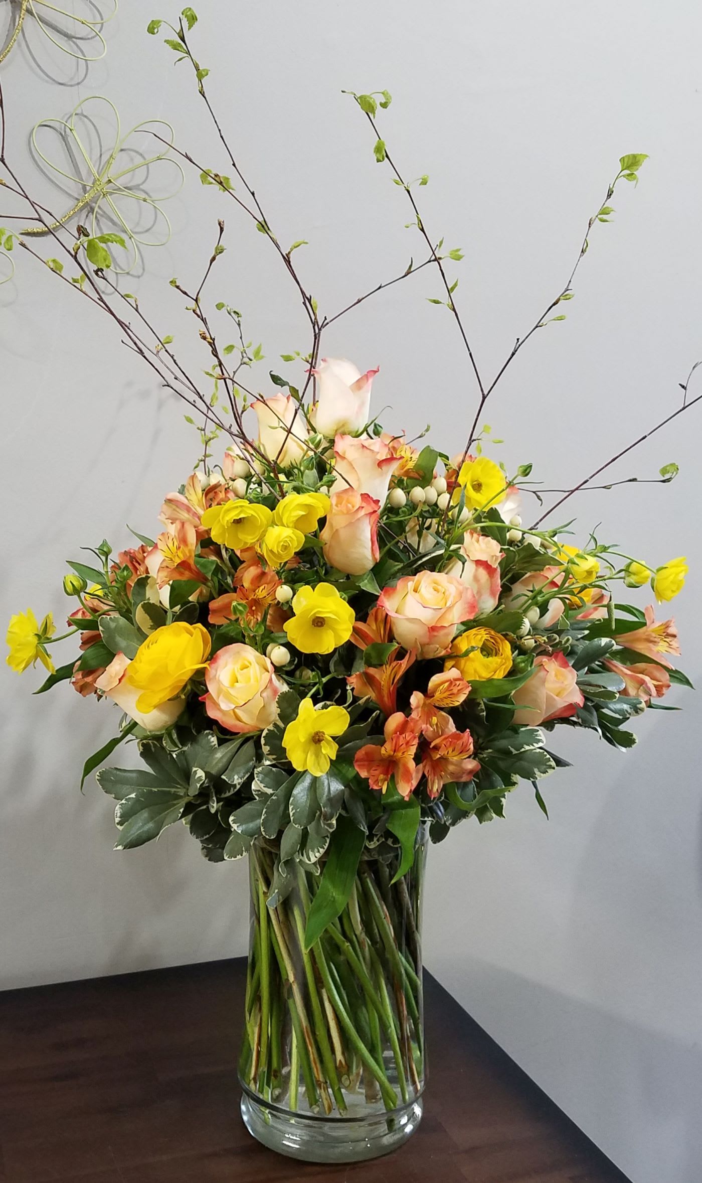 Tall vase arrangement with peach roses, yellow ranunculus, and orange alstroemeria accented by airy branches.