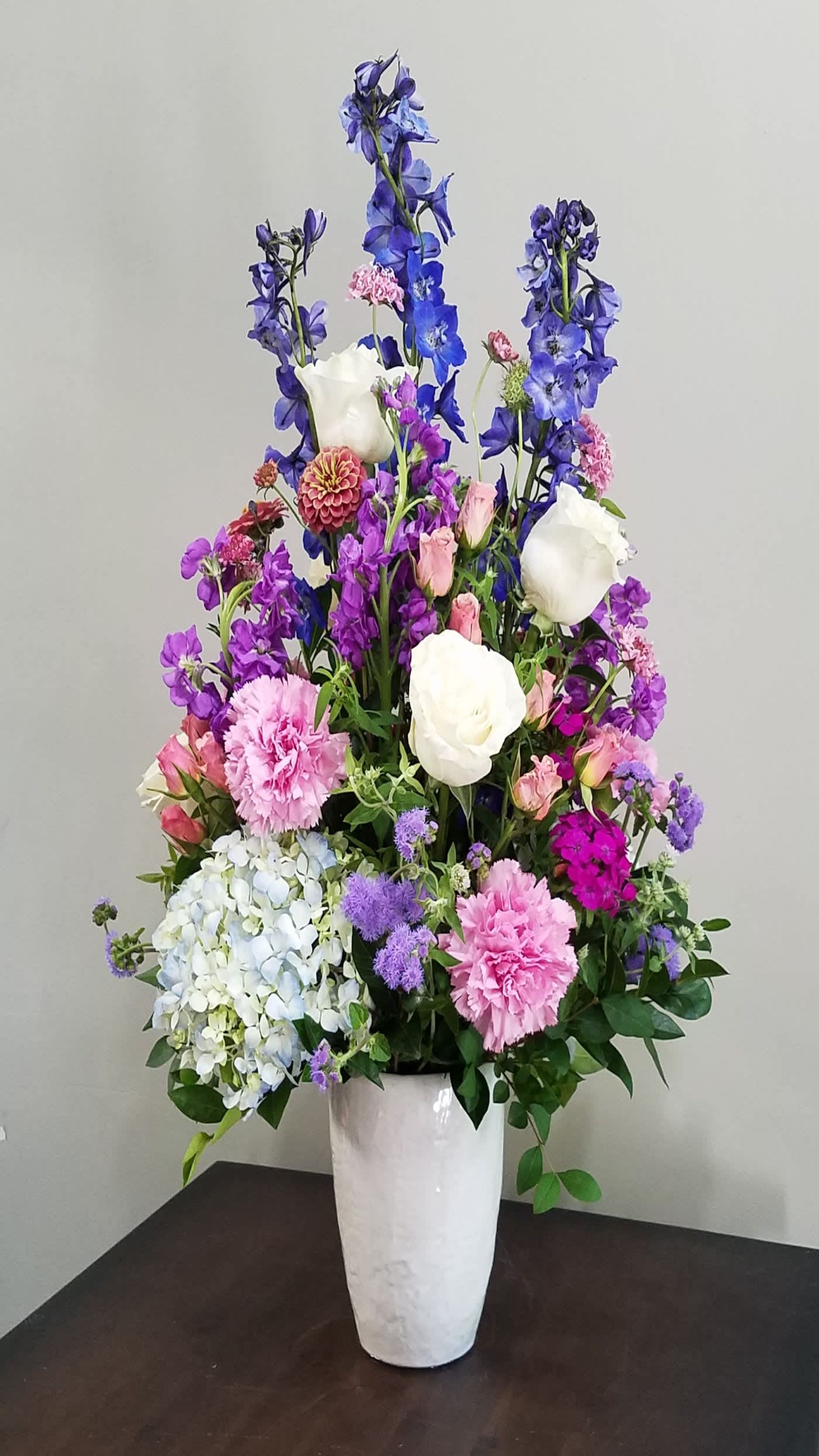 Tall mixed arrangement of purple, pink, blue, and white flowers in a white vase on a dark table