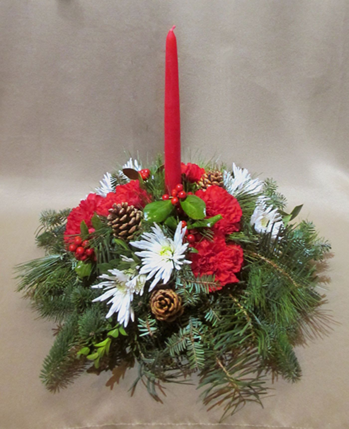 Low centerpiece of red carnations, white mums, evergreen branches and a tall red taper candle.