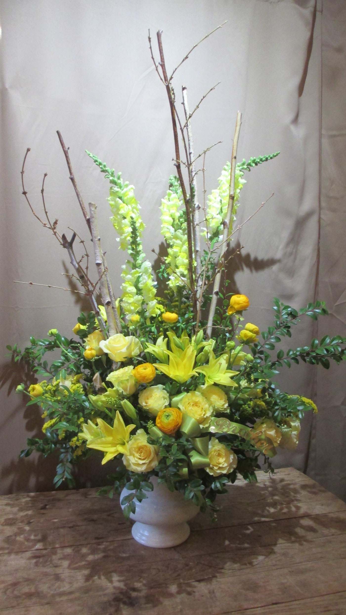 Tall yellow flower arrangement with lilies, roses, and branches in a white urn vase