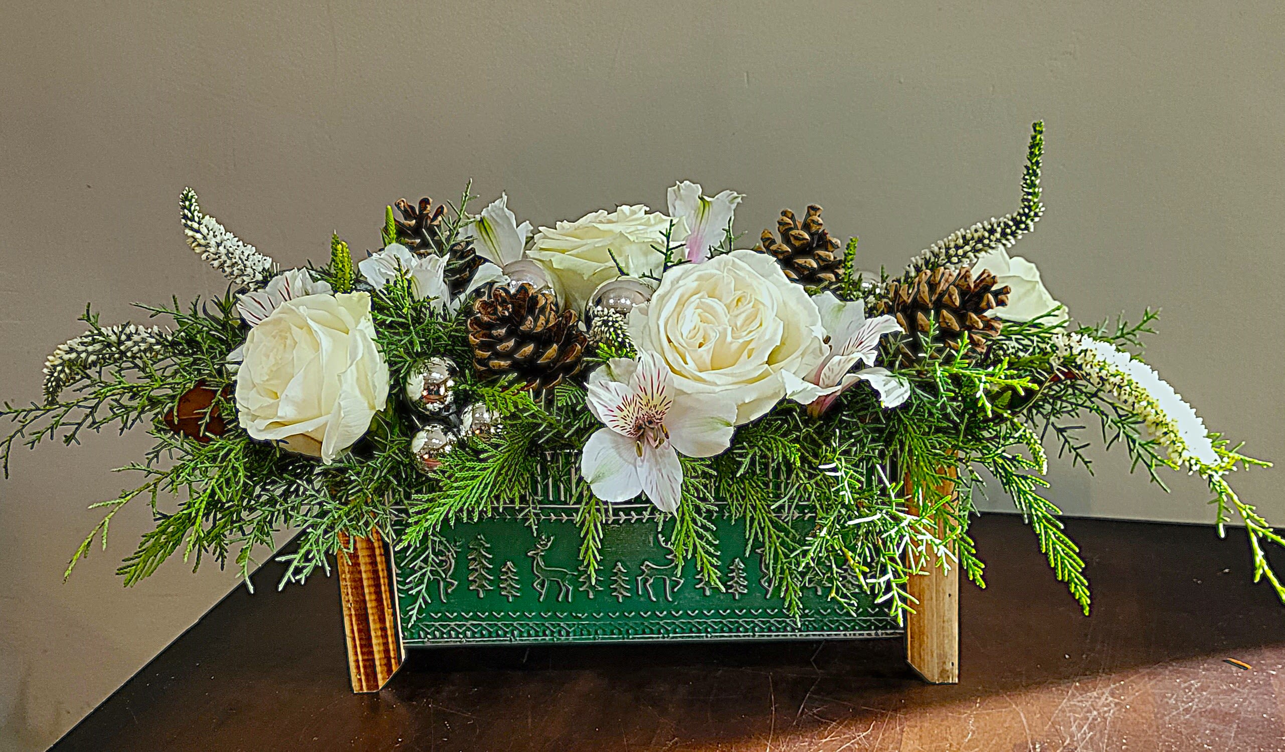 Low rectangular holiday arrangement with white flowers, pinecones, and evergreens in a green wooden box.