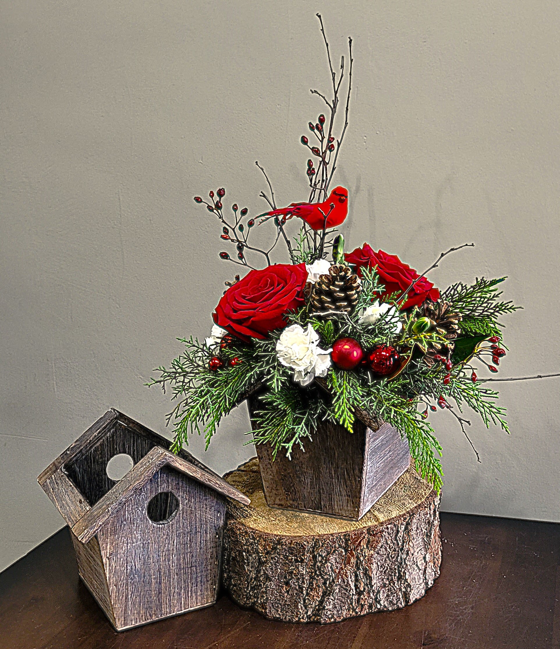 Rustic birdhouse arrangement with red roses, white flowers, pinecones and a red cardinal on a wood slice base.