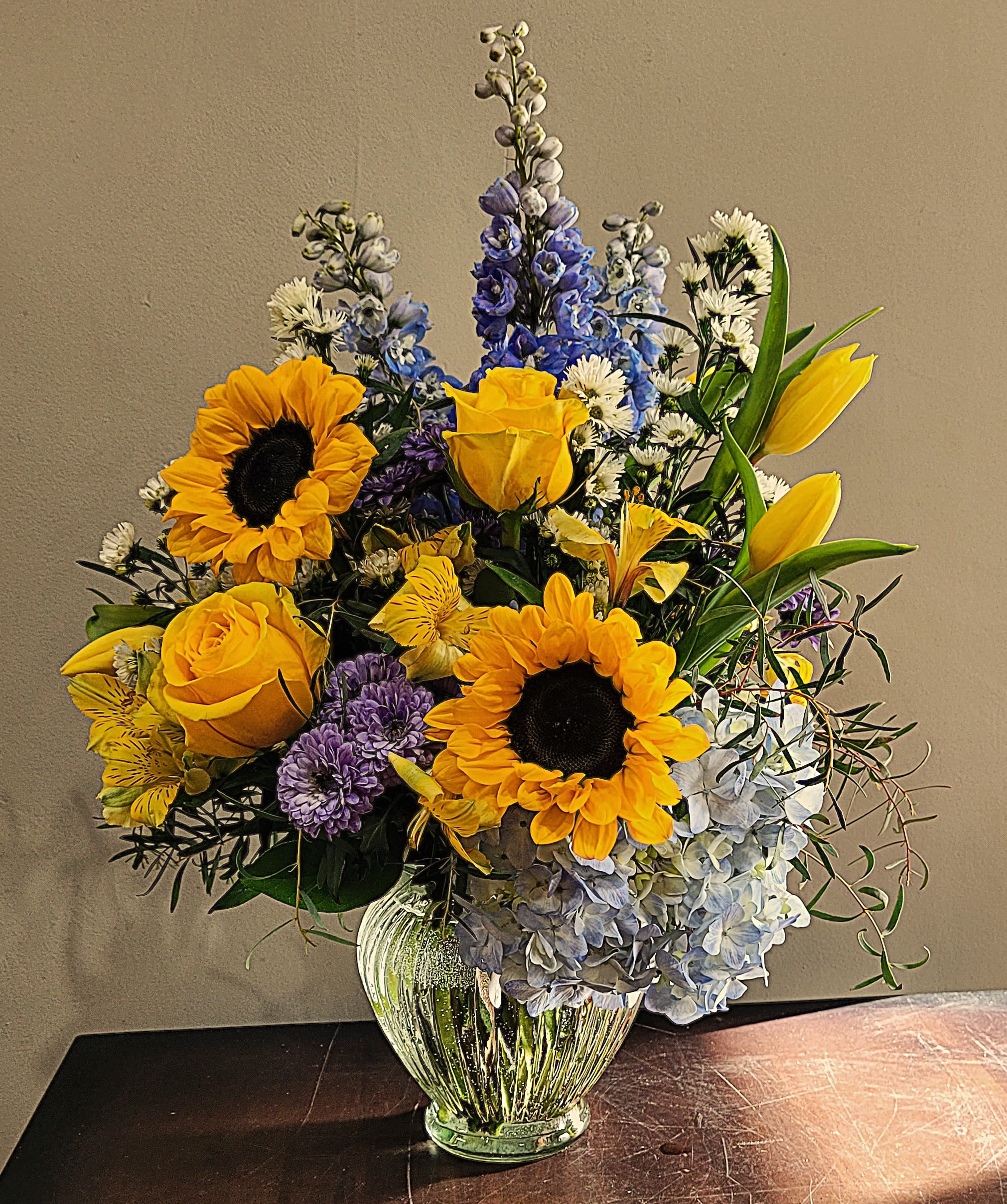 Arrangement of yellow sunflowers, roses, and blue hydrangeas in a clear glass vase