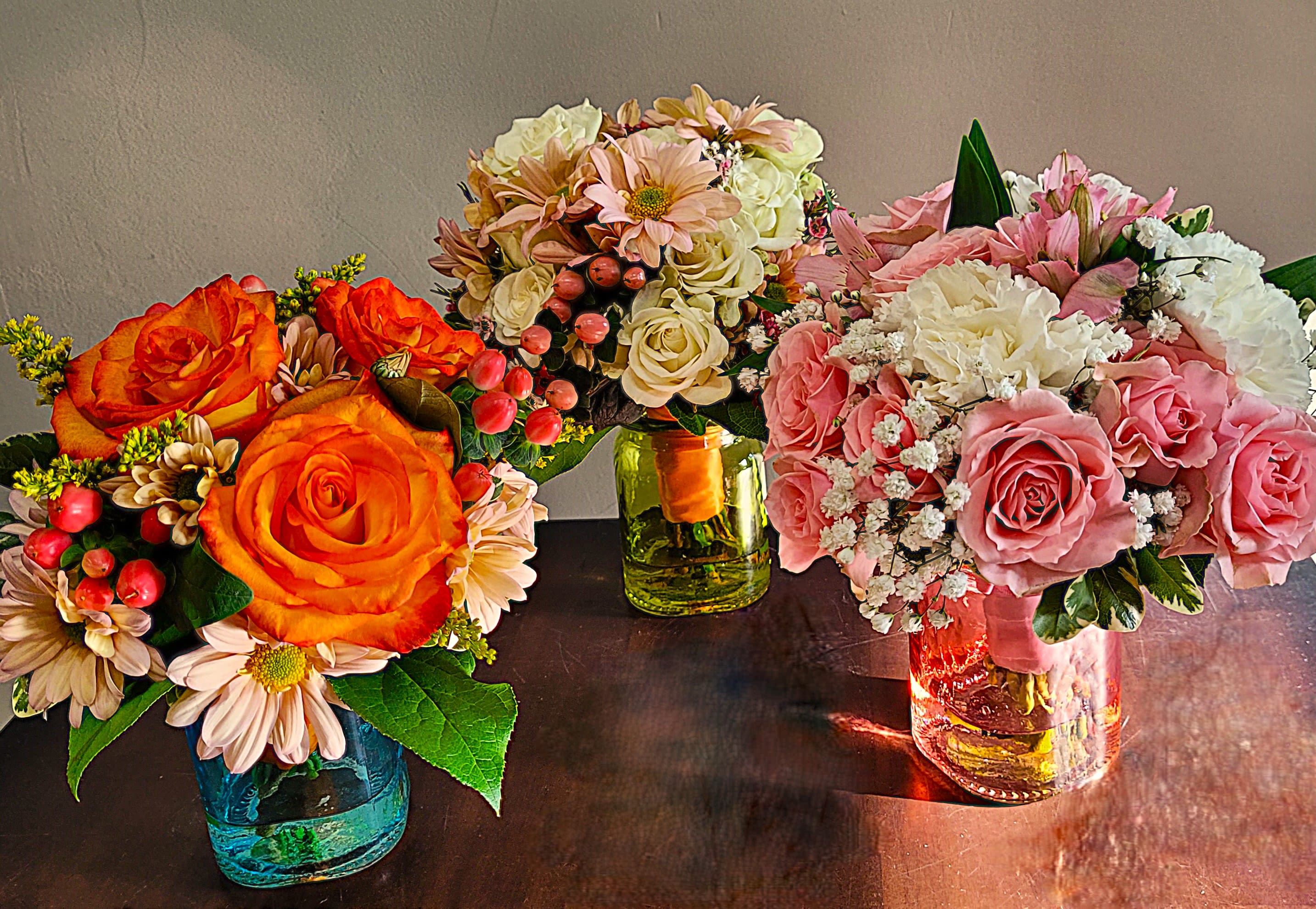 Three small mixed flower arrangements with roses and daisies in colored glass jars on a table.