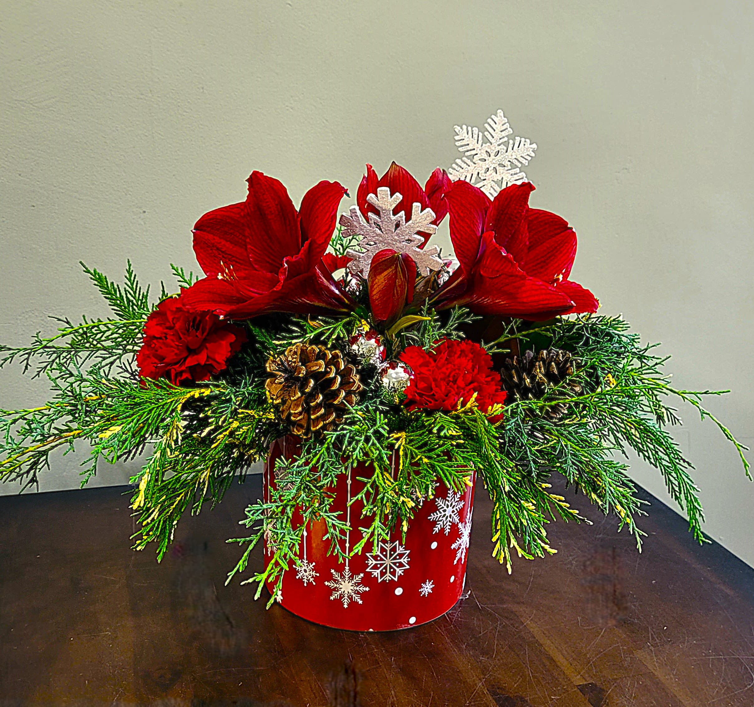 Low red holiday arrangement with red amaryllis, red carnations, evergreens, and pine cones in a snowflake container.