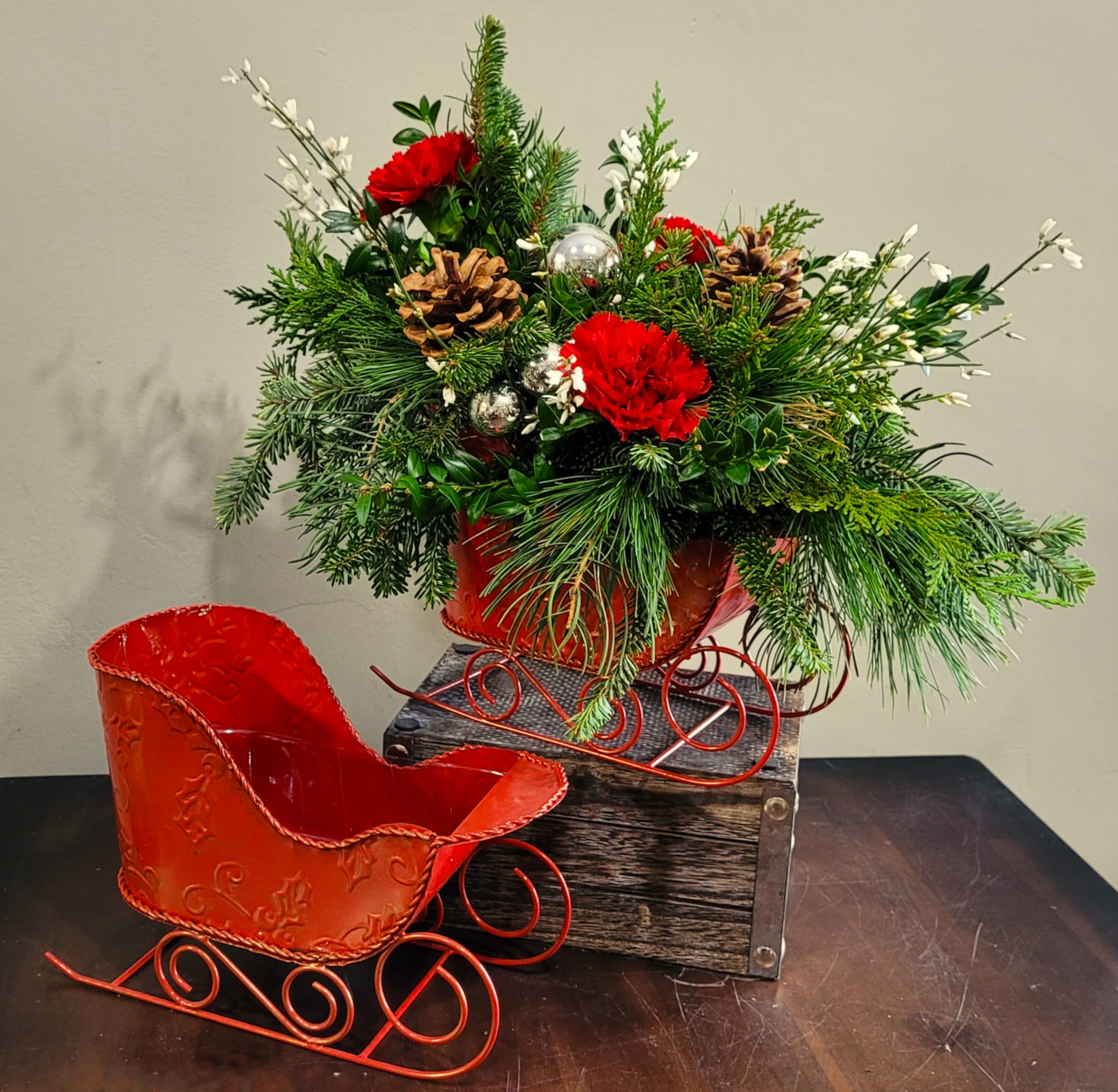 Red carnations and pinecones in a red metal sleigh with evergreens and silver ornaments beside an empty matching sleigh.
