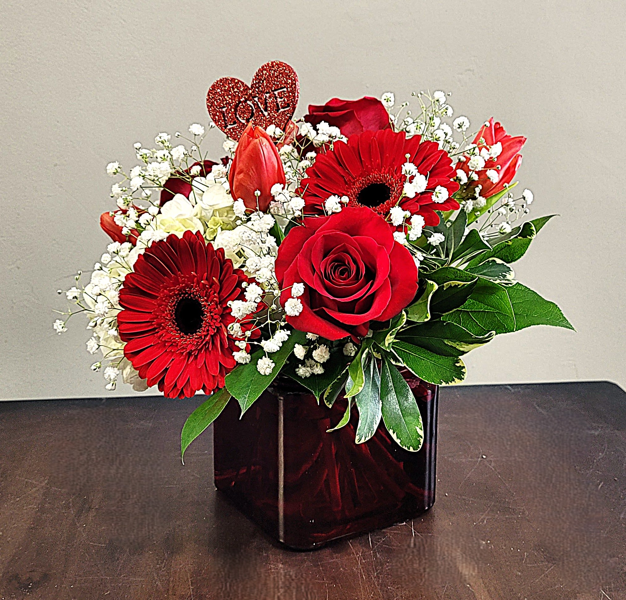 Red roses, gerbera daisies, and tulips with white accents in a red glass cube vase and heart LOVE pick