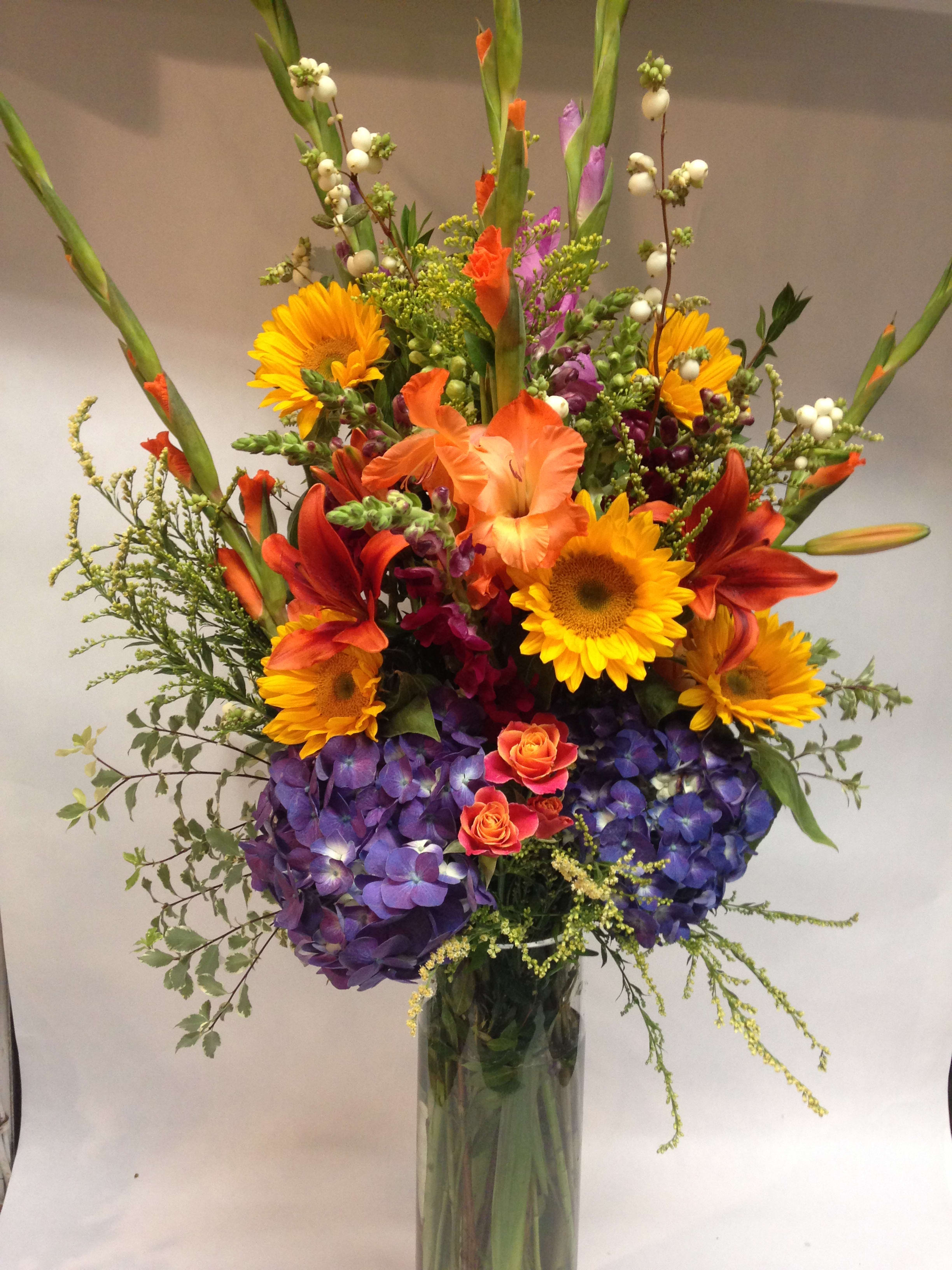 Tall mixed bouquet with sunflowers, lilies, and purple hydrangeas in a glass vase