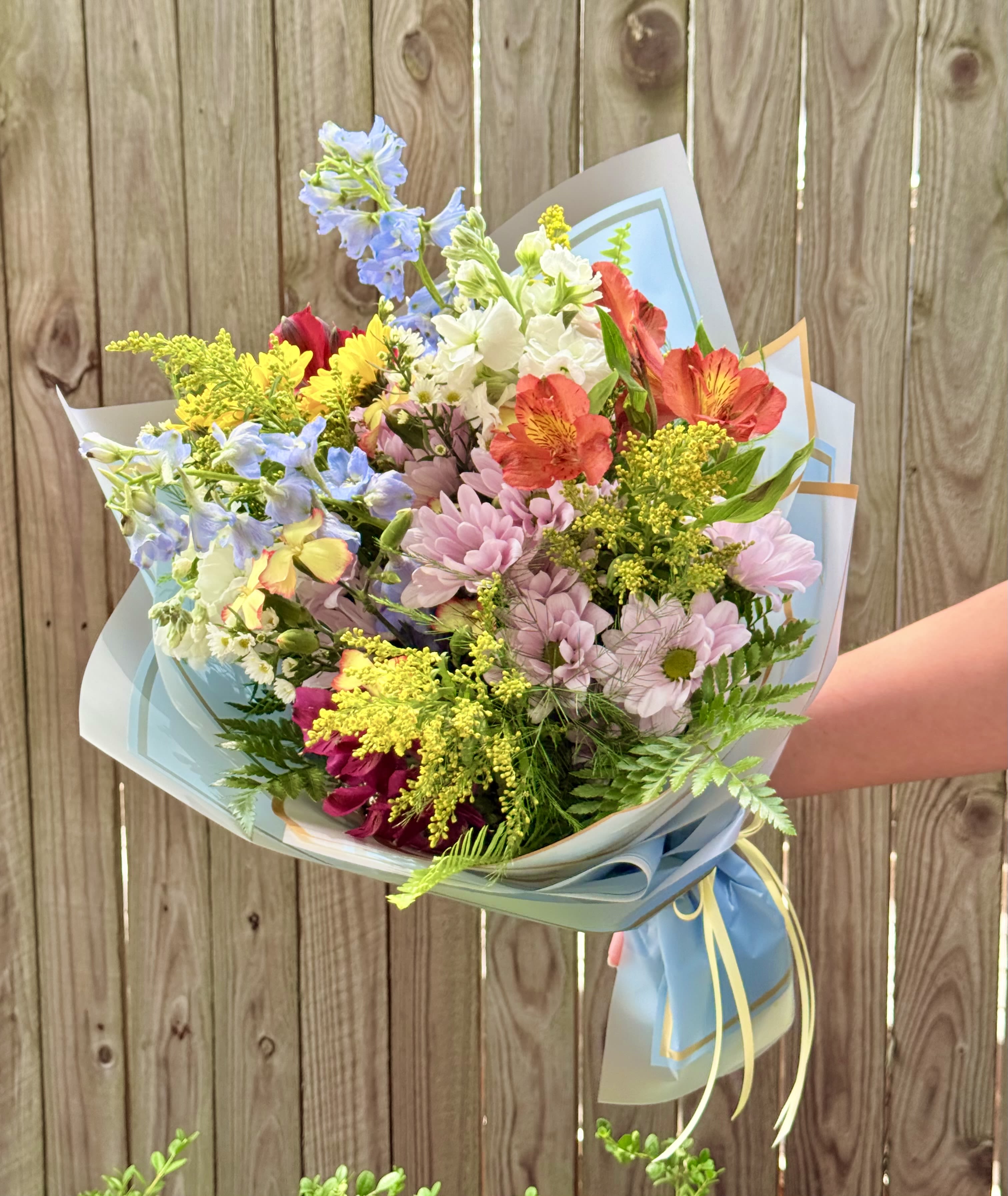 Barefoot Summer Wrap - A cheerful garden-style bouquet that brightens any celebration - perfect as a birthday bouquet, thank-you gift, or just-because surprise. Soft pastels mingle with lively pops of color so the person you're thinking of will feel genuinely noticed: lavender daisies and orange alstroemeria create warmth, while pale blue delphinium spikes and sunny yellow filler bring playful contrast.  This mixed bouquet is wrapped in layered light blue and white decorative paper and finished with a ribbon tie for a gift-ready presentation. The texture ranges from the daisy centers' soft faces to the alstroemerias' delicate petals and the delphiniums' vertical accents, giving the arrangement a whimsical, hand-picked look that sits nicely in a vase once unwrapped. Ideal for cheering a friend, celebrating a milestone, or sending a bright thinking-of-you gesture.