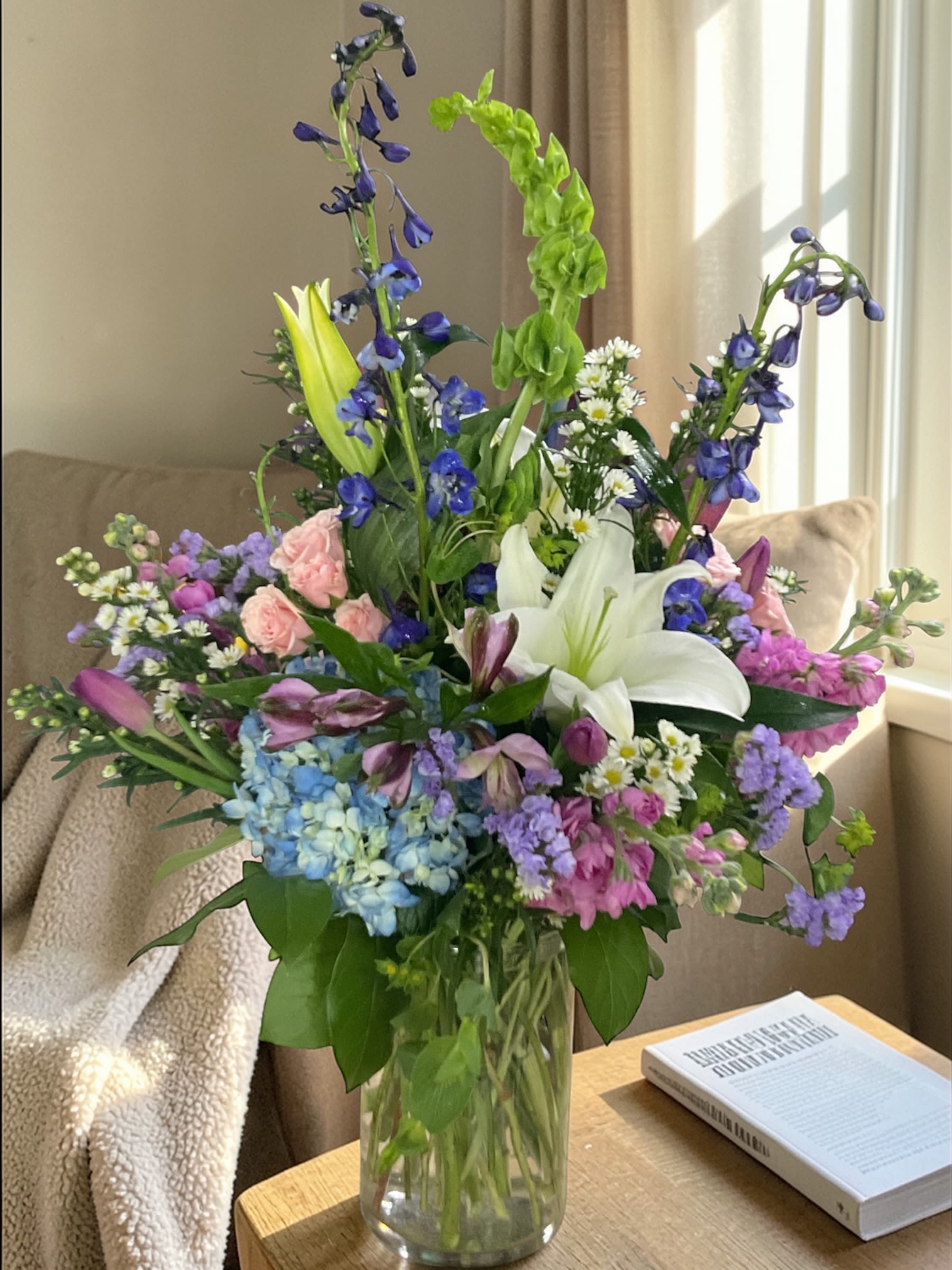 Garden Daydream  - Garden Daydream is soft, colorful, and full of relaxed garden charm. A blend of seasonal blooms in shades of blue, pink, lavender, and fresh green comes together with lilies, hydrangea, and delphinium for a design that feels natural, airy, and easy.  Arranged in a clear glass vase, the look leans into that “just gathered” feel—loose, balanced, and full without being heavy or overworked.  A beautiful choice for birthdays, celebrations, encouragement, thinking of you, or simply sending something fresh and uplifting.  Each arrangement is designed with the freshest stems available. Varieties may vary slightly based on availability while maintaining the same color palette and overall garden style.  Available for local flower delivery through Aspen Tower Florist, serving Greensburg and Shelbyville along with Rushville, Batesville, Westport, Oldenburg, St. Paul, Waldron, and Milroy.
