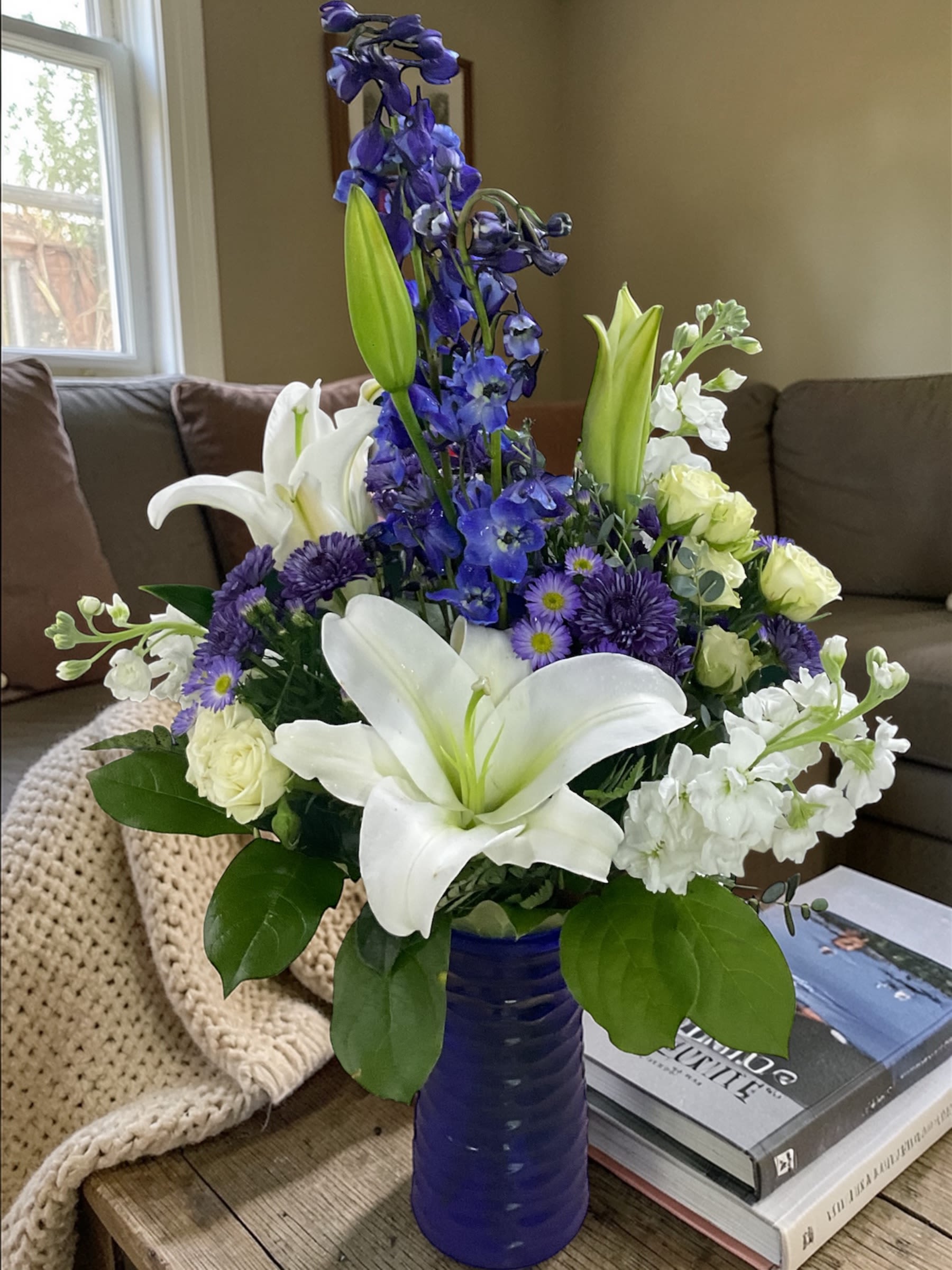 River Breeze - River Breeze is fresh, cool, and effortlessly elegant. Graceful white lilies and rich blue delphinium are paired with soft seasonal blooms, all arranged in a striking cobalt vase for a design that feels calm, balanced, and visually refreshing.  The flowing mix of crisp whites and deep blues creates a clean, composed look inspired by the quiet movement of water—light, open, and easy on the eye without feeling overworked.  A beautiful choice for birthdays, celebrations, encouragement, sympathy, thinking of you, or sending something peaceful and refined.  Each arrangement is designed with the freshest stems available. Varieties may vary slightly based on availability while maintaining the same blue and white palette and overall style.  Available for local flower delivery through Aspen Tower Florist, serving Greensburg and Shelbyville along with Rushville, Batesville, Westport, Oldenburg, St. Paul, Waldron, Milroy, and Hope.