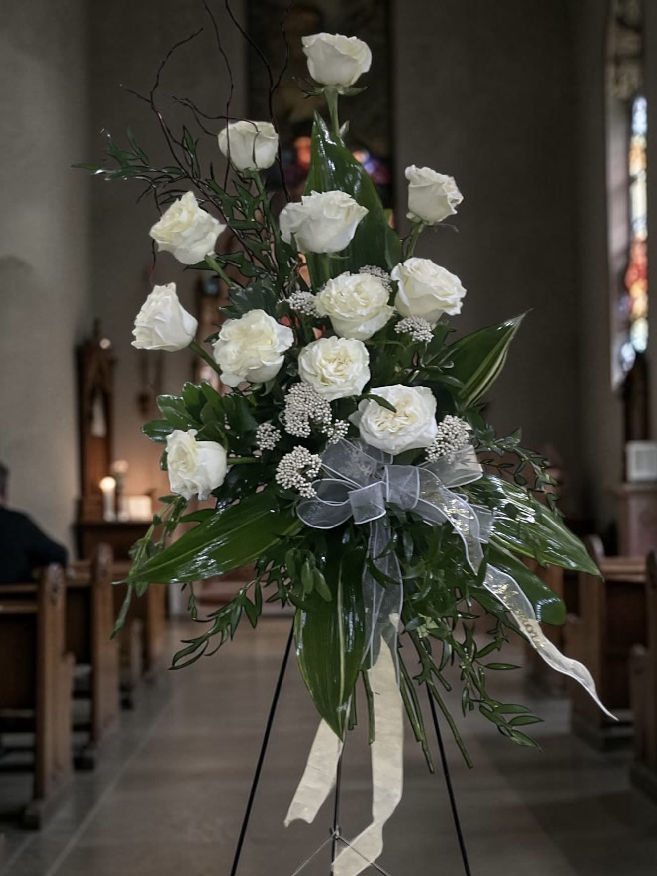 Forever Remembered Spray on Easel - When words are difficult to find, Forever Remembered offers a timeless expression of love, respect, and remembrance. Featuring elegant white roses arranged on a standing easel with lush greenery and classic accents, this sympathy tribute creates a beautiful focal display for funeral services, memorial services, visitations, celebration of life gatherings, churches, and graveside services.  The Standard design includes 12 white roses. Deluxe features 18 roses for greater fullness and presence. Premium showcases 24 roses for a more abundant and dramatic display.  White roses have long symbolized honor, devotion, reverence, innocence, and enduring love, making them one of the most requested flowers for funeral and memorial arrangements. Whether honoring a parent, spouse, grandparent, sibling, veteran, friend, or loved one, this standing spray delivers a heartfelt message of sympathy and support.  Designed for delivery to funeral homes, churches, memorial chapels, and graveside services, Forever Remembered provides an elegant way to celebrate a life, express condolences, and share lasting memories.
