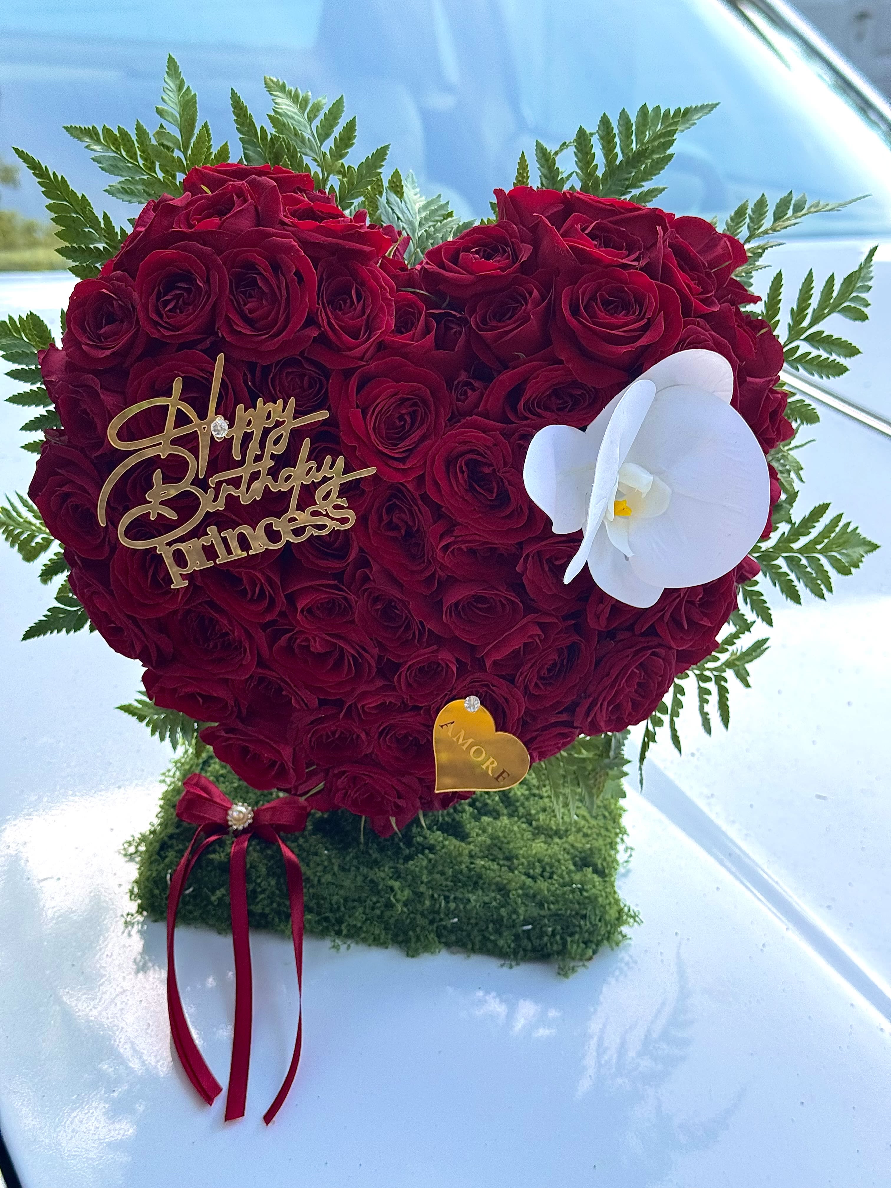 Luxury 3D Heart Floral Arrangement - A romantic 3D heart-shaped floral sculpture designed to make a bold and unforgettable statement.  This handcrafted design is created with fresh premium red roses, carefully arranged to form a full dimensional heart that stands beautifully from every angle. Accented with elegant white orchid blooms, this piece symbolizes passion, deep love, and timeless romance.  Its sculpted 3D structure creates visual impact while maintaining refined elegance — making it perfect for anniversaries, romantic surprises, proposals, Valentine’s Day, birthdays, or any meaningful celebration.  Each arrangement is thoughtfully designed with attention to detail, ensuring a luxurious presentation that reflects the signature style of AMORE.  Please note: Orchid accents and flower tones may vary slightly depending on seasonal availability while preserving the overall elegant aesthetic.  This is not just a bouquet — it is a floral statement piece.