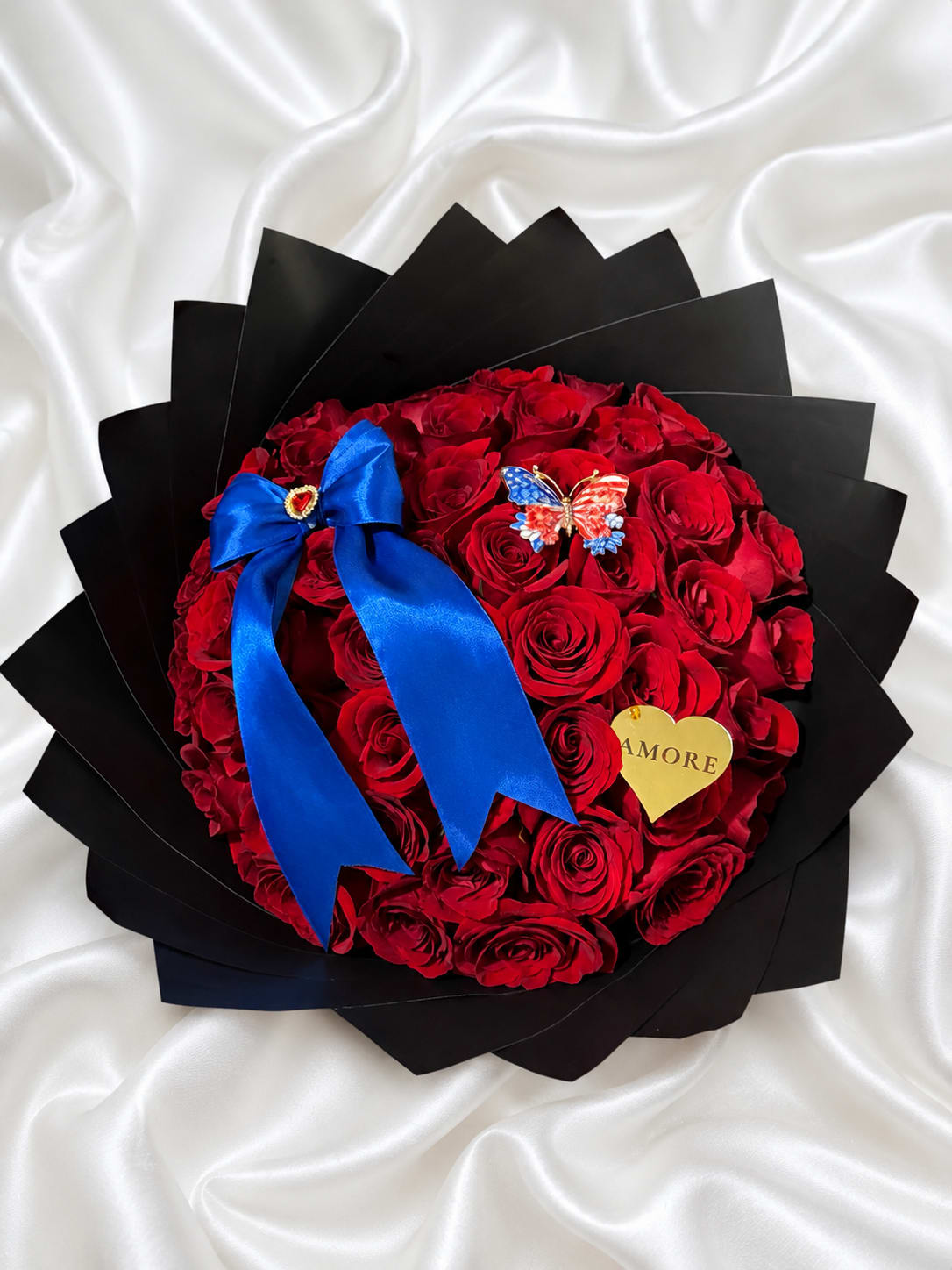     4th of July Rose Heart with Patriotic Butterfly - Celebrate freedom, love, and unforgettable moments with our Patriotic Red Rose Heart with Butterfly Brooch. This elegant arrangement features premium fresh red roses, luxurious black wrapping, and a royal blue satin ribbon for a sophisticated patriotic look.  The highlight of this design is its beautiful patriotic butterfly brooch, which is removable and can also be worn as a clothing accessory, making this gift a lasting keepsake even after the flowers have been enjoyed.  Perfect for Independence Day, birthdays, anniversaries, military celebrations, or simply surprising someone special, each arrangement is handcrafted to order by AMORE Flowers using carefully selected premium roses to ensure exceptional beauty and freshness.  Available in 25, 50, or 75 roses for every occasion.