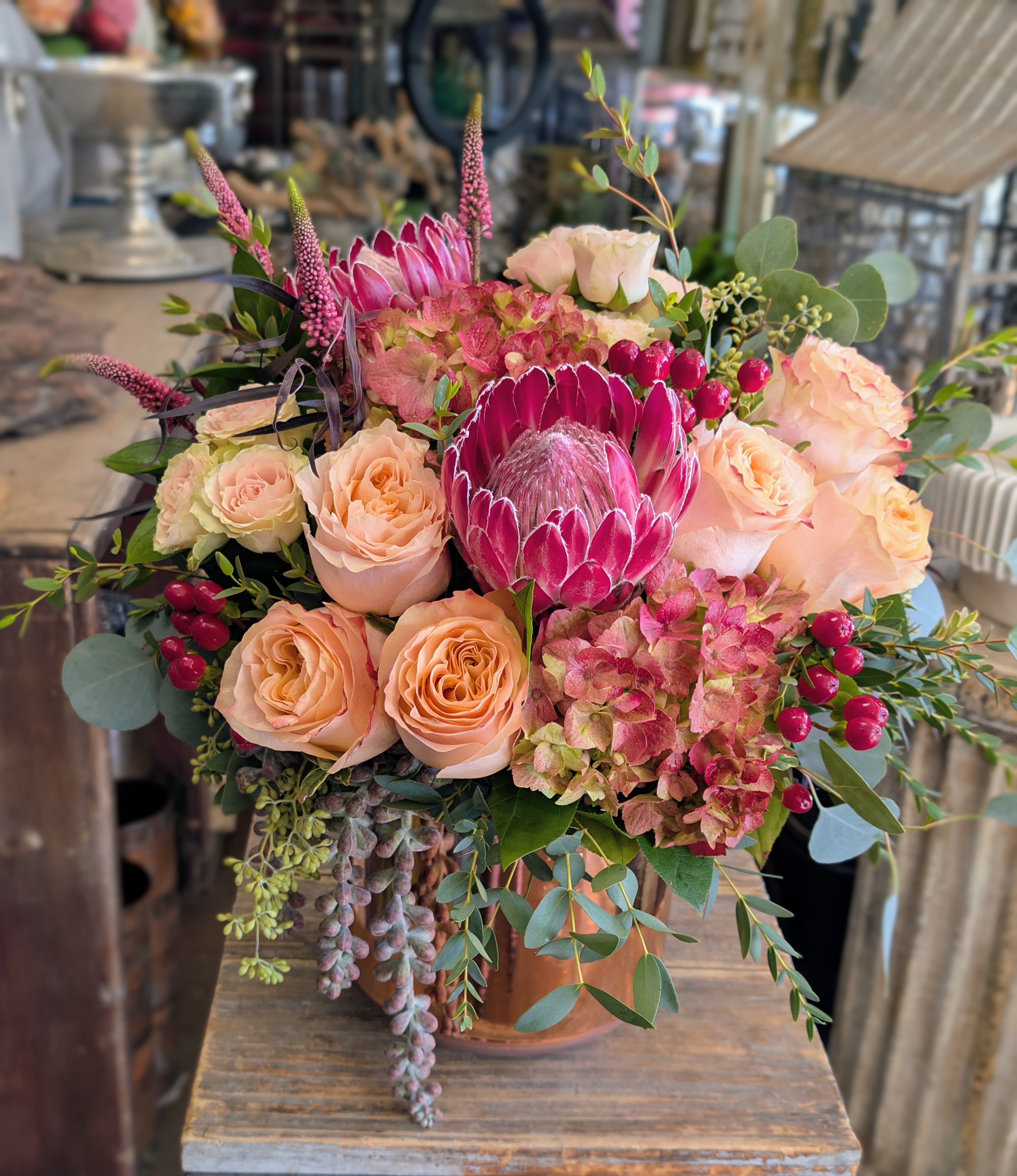 Bordeaux - Bordeaux is a rich, romantic statement arrangement inspired by old-world wine tones and modern desert luxury. Featuring dramatic protea as the centerpiece, Bordeaux is layered with blush and peach garden roses, antique hydrangea, hypericum berries, textural veronica, and cascading succulent accents, all finished with lush eucalyptus and mixed greenery. Designed in a warm copper vessel, this arrangement blends deep berry hues with soft champagne florals for a look that feels both bold and refined.  Handcrafted by Windmill Floral Expressions, Bordeaux is perfect for anniversaries, celebrations, romantic gestures, or anyone drawn to moody, European-inspired florals. Each arrangement is custom designed with premium blooms and thoughtful detail, making every piece truly one of a kind.  Based in Las Vegas and Henderson, Windmill Floral Expressions specializes in bespoke floral designs, luxury arrangements, and curated botanical moments. Same-day delivery is available throughout Las Vegas, Henderson, Anthem, Inspirada, MacDonald Ranch, Ascaya, and surrounding areas.  Let Bordeaux set the mood — dramatic, elegant, and unforgettable.