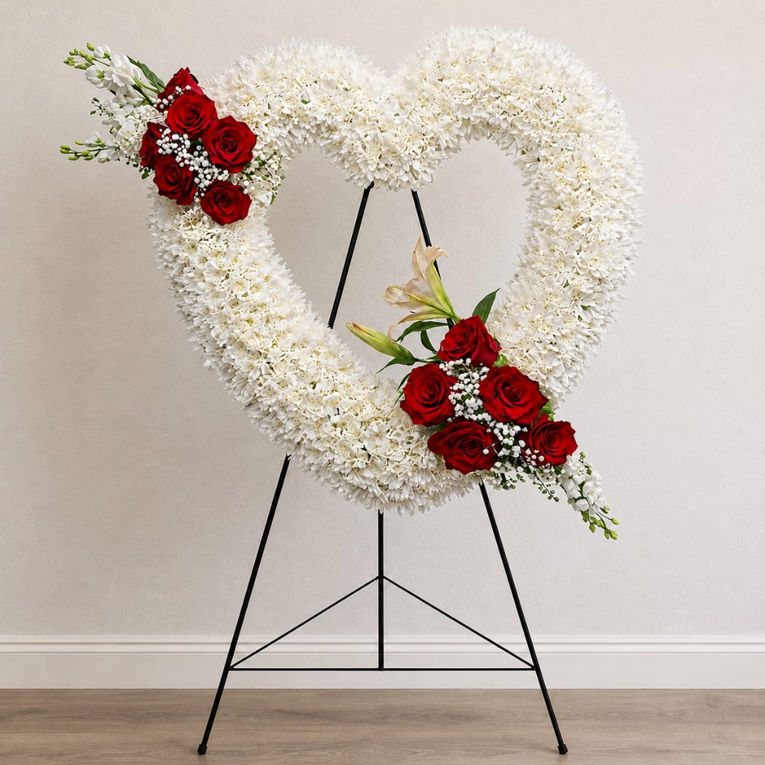 Heart-shaped white floral wreath with red rose clusters on a black stand