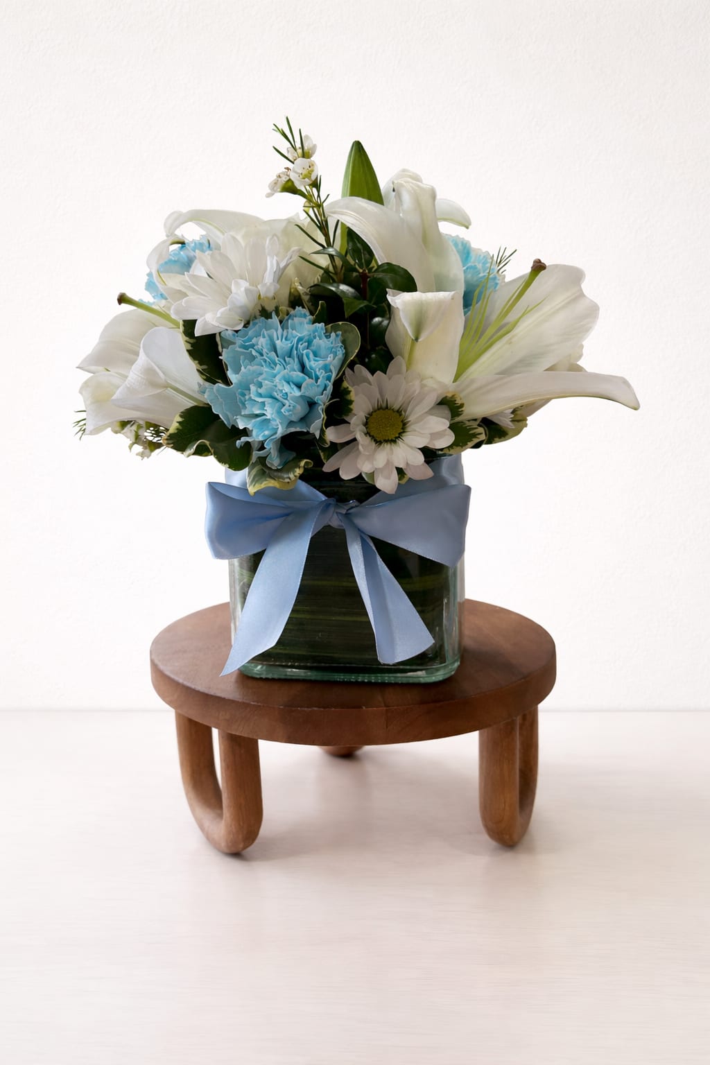 White lilies and daisies with blue carnations in a glass vase with a blue ribbon