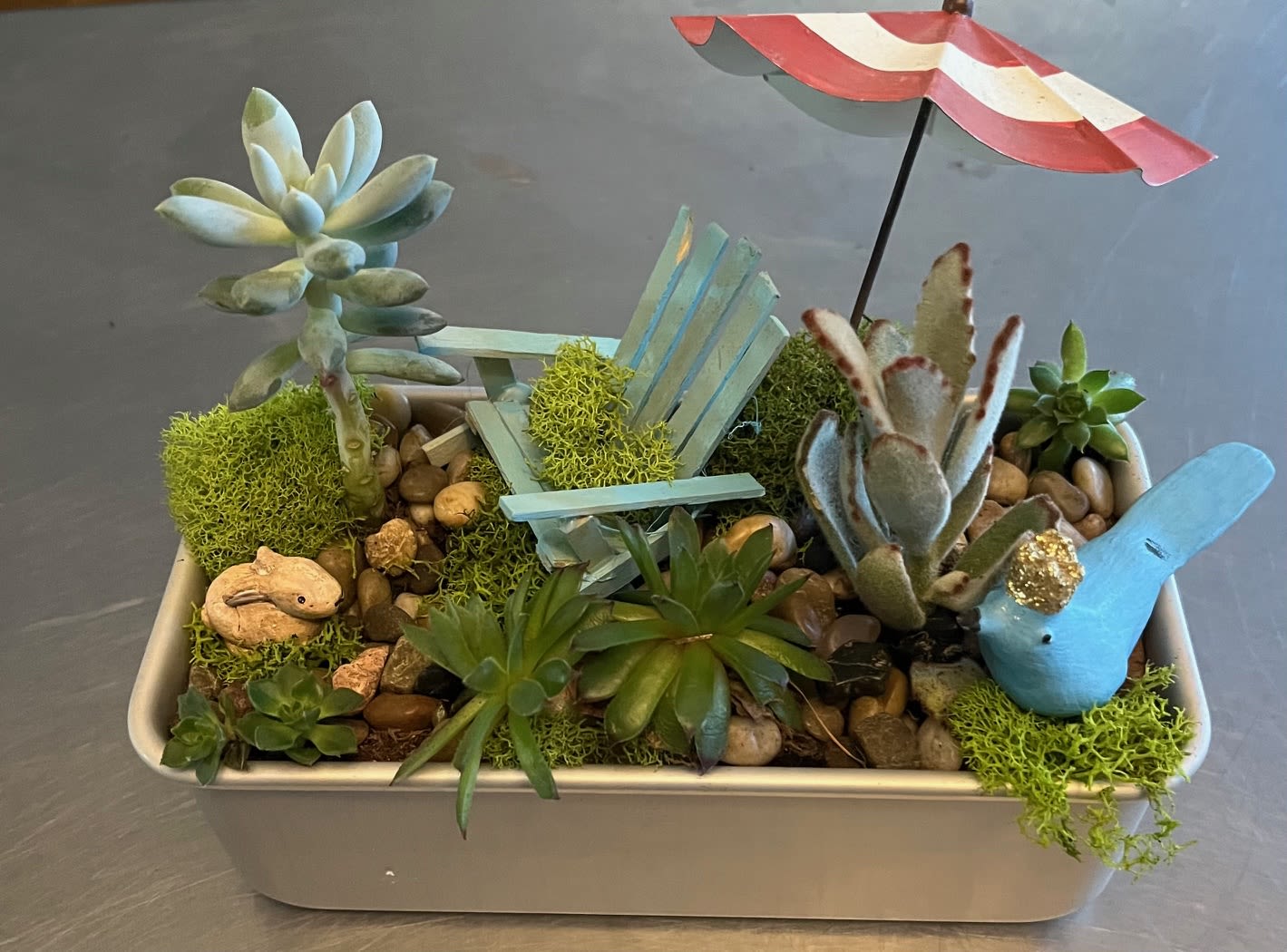 Succulent arrangement in a rectangular planter with a tiny chair and umbrella