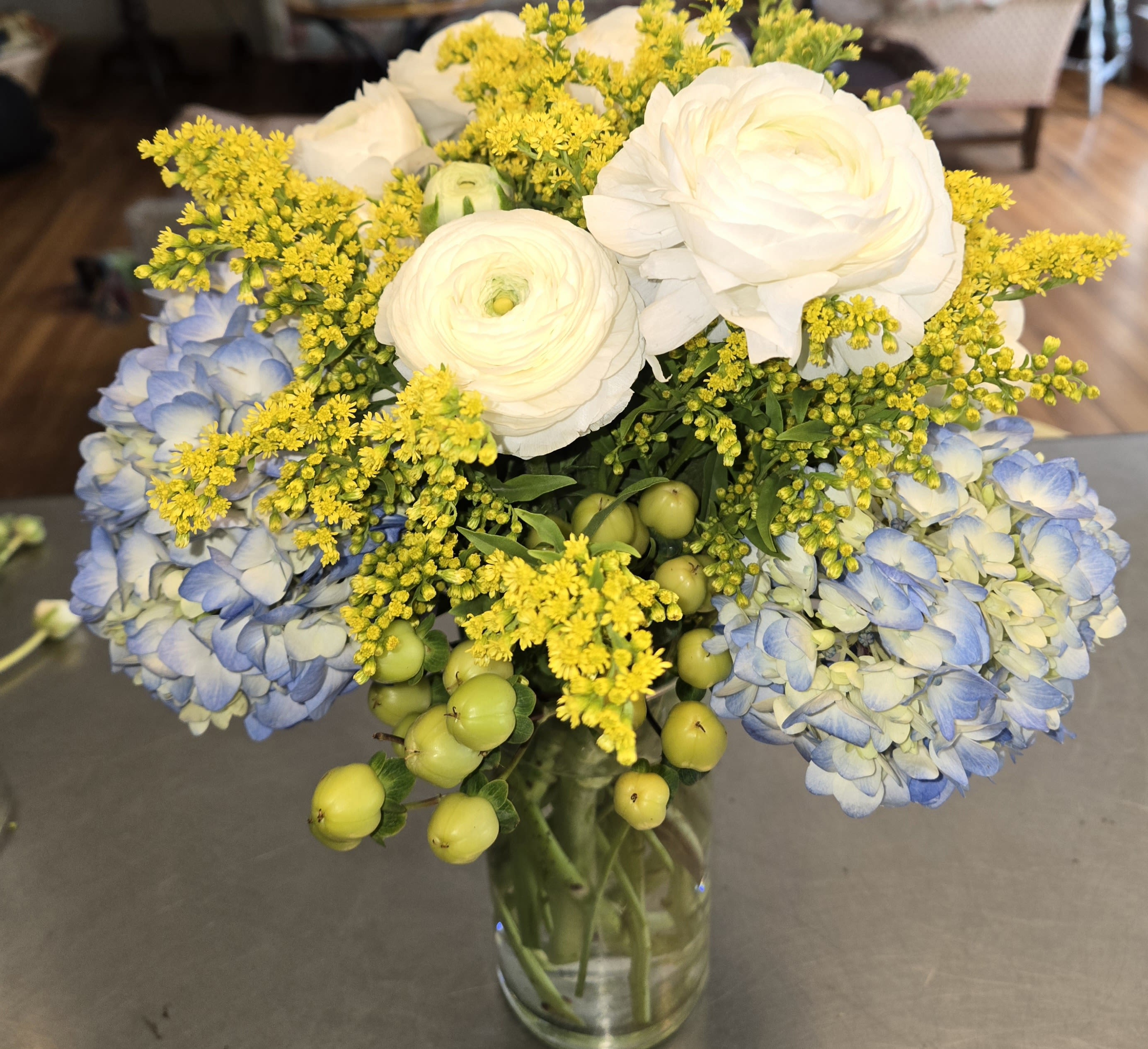 Bouquet of white ranunculus, blue hydrangea, and yellow filler in a glass vase