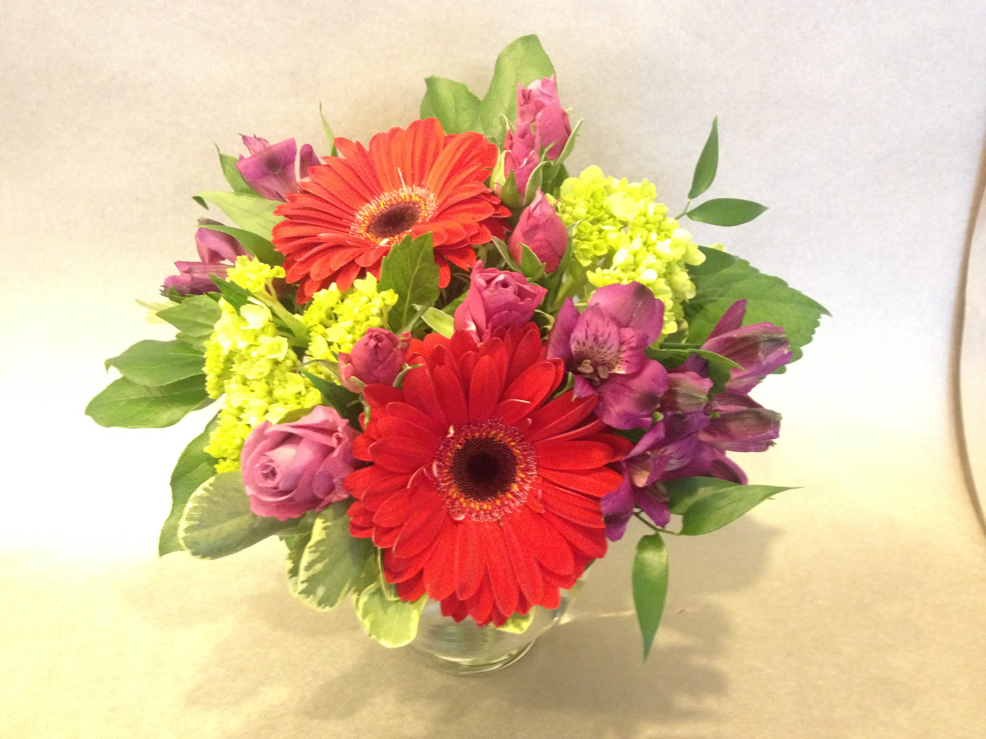 Bouquet of red gerbera daisies, pink roses, and purple flowers in a glass vase
