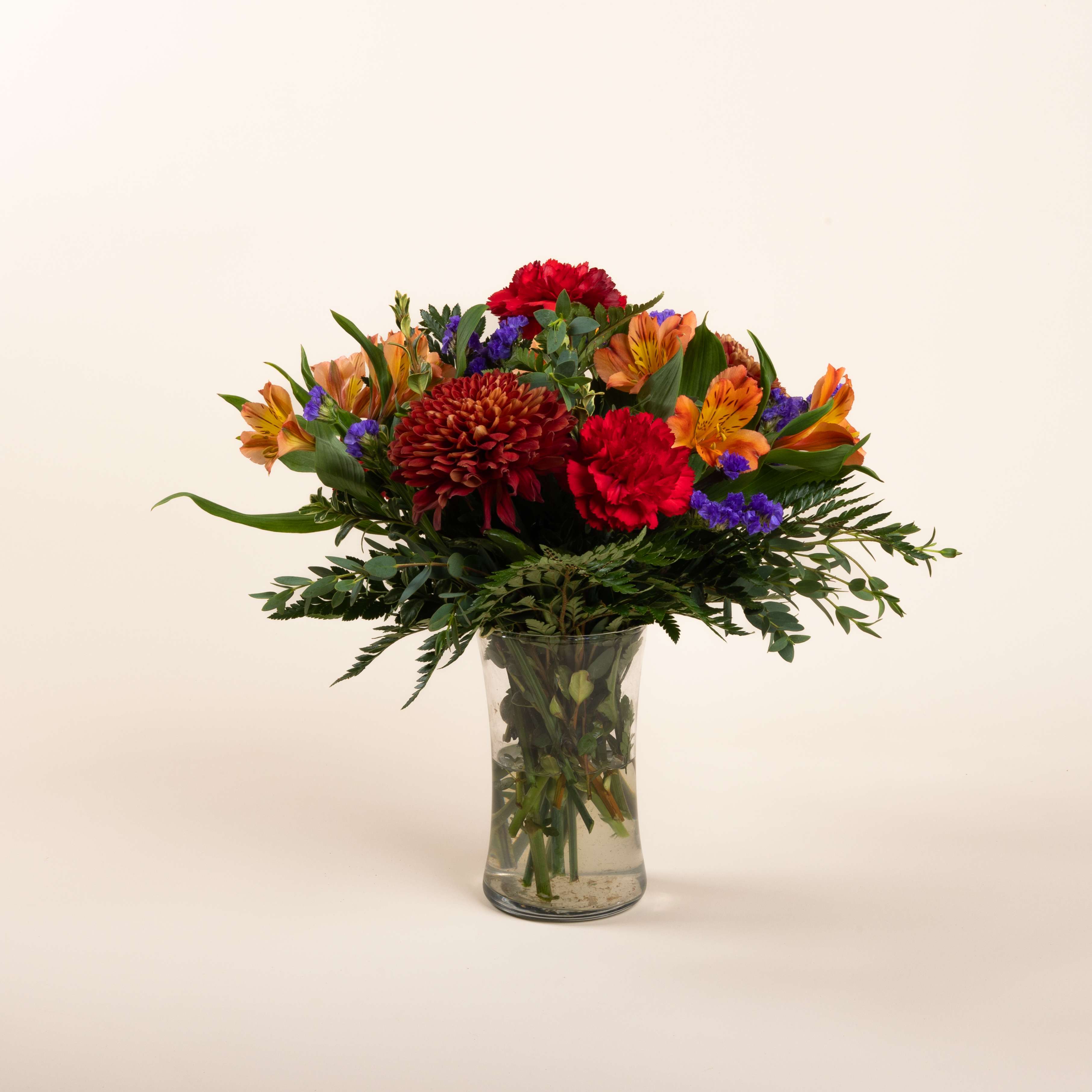 Woodsy - Bronze Cremona, red carnations, orange alstromeria, and purple statice with leather leaf, gunni eucalyptus, and oregonia designed in a clear vase.