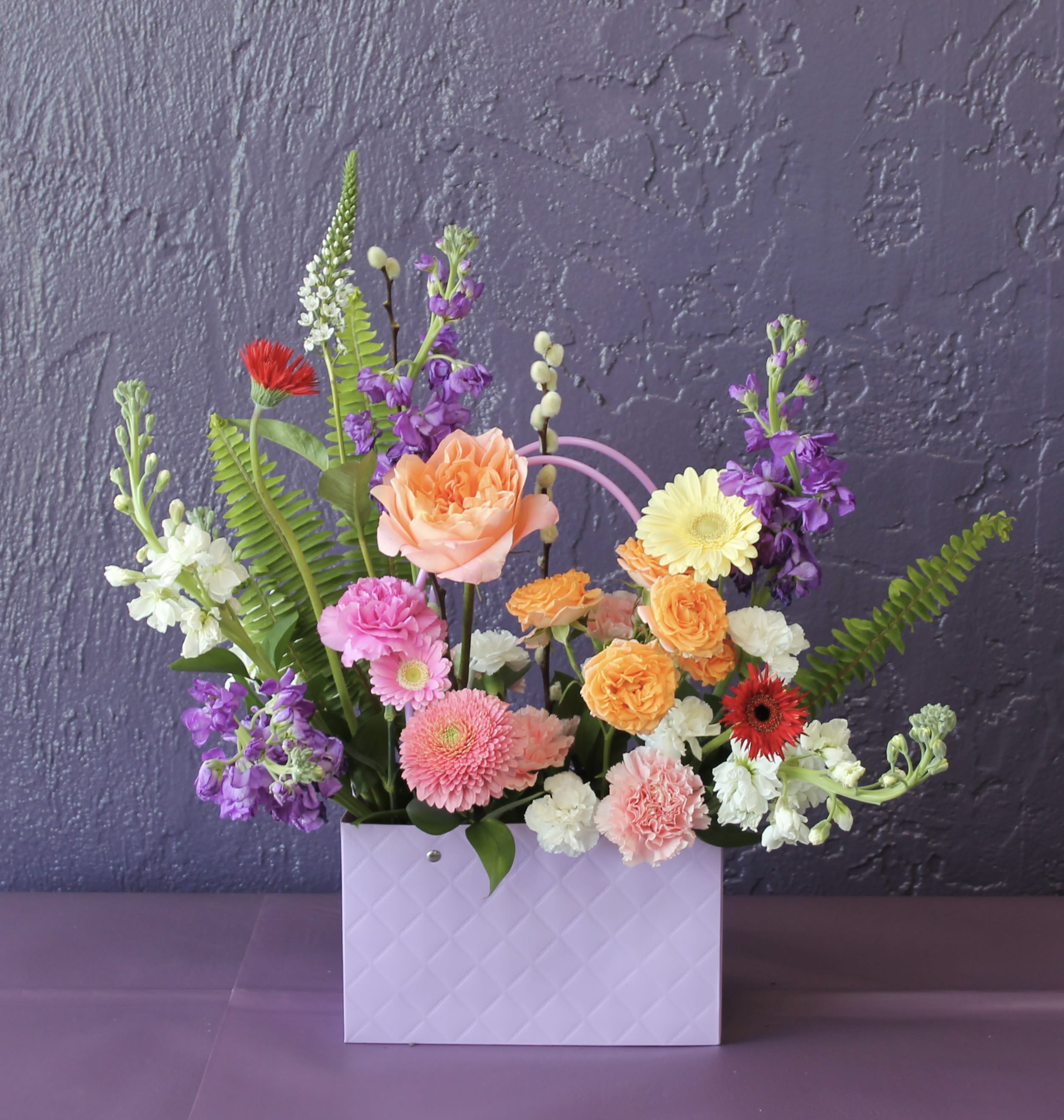 Colorful mixed flower arrangement in a lavender gift bag-style container against a purple wall